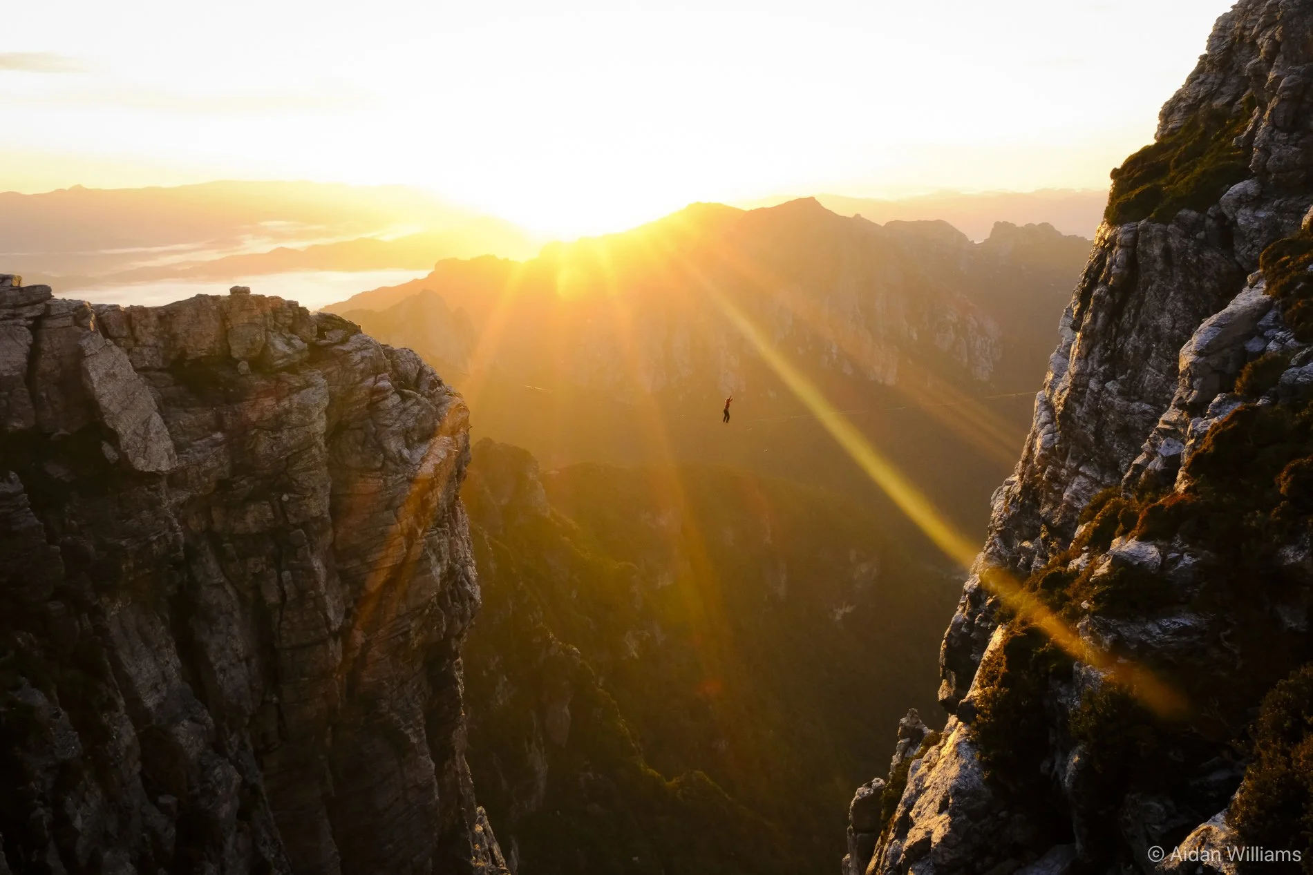 Person hanging suspended between two high mountain cliffs during a sunrise with sun rays shining through clouds.