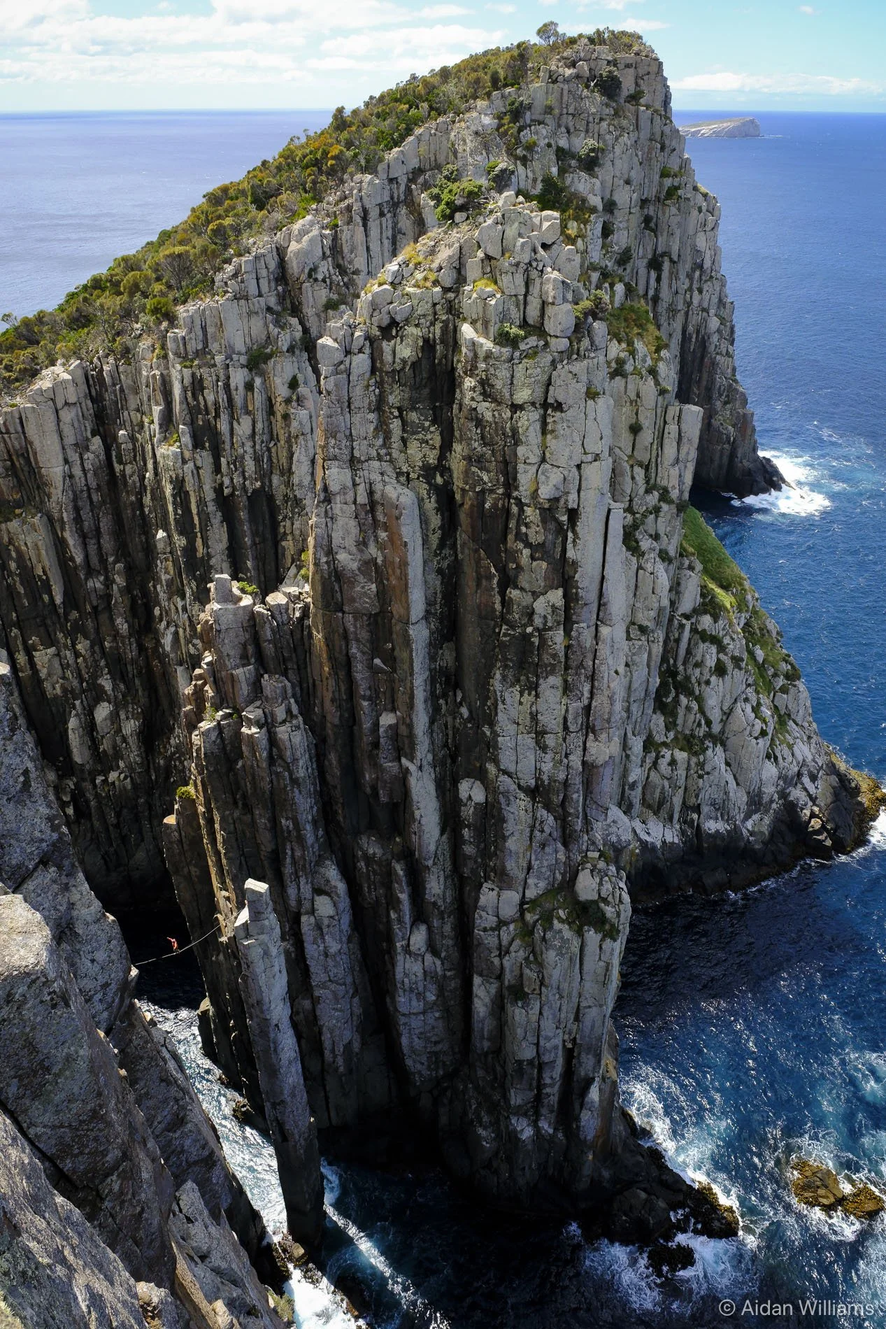 Cliffside with tall, vertical rock formations extending into the ocean, surrounded by blue water and greenery on top.