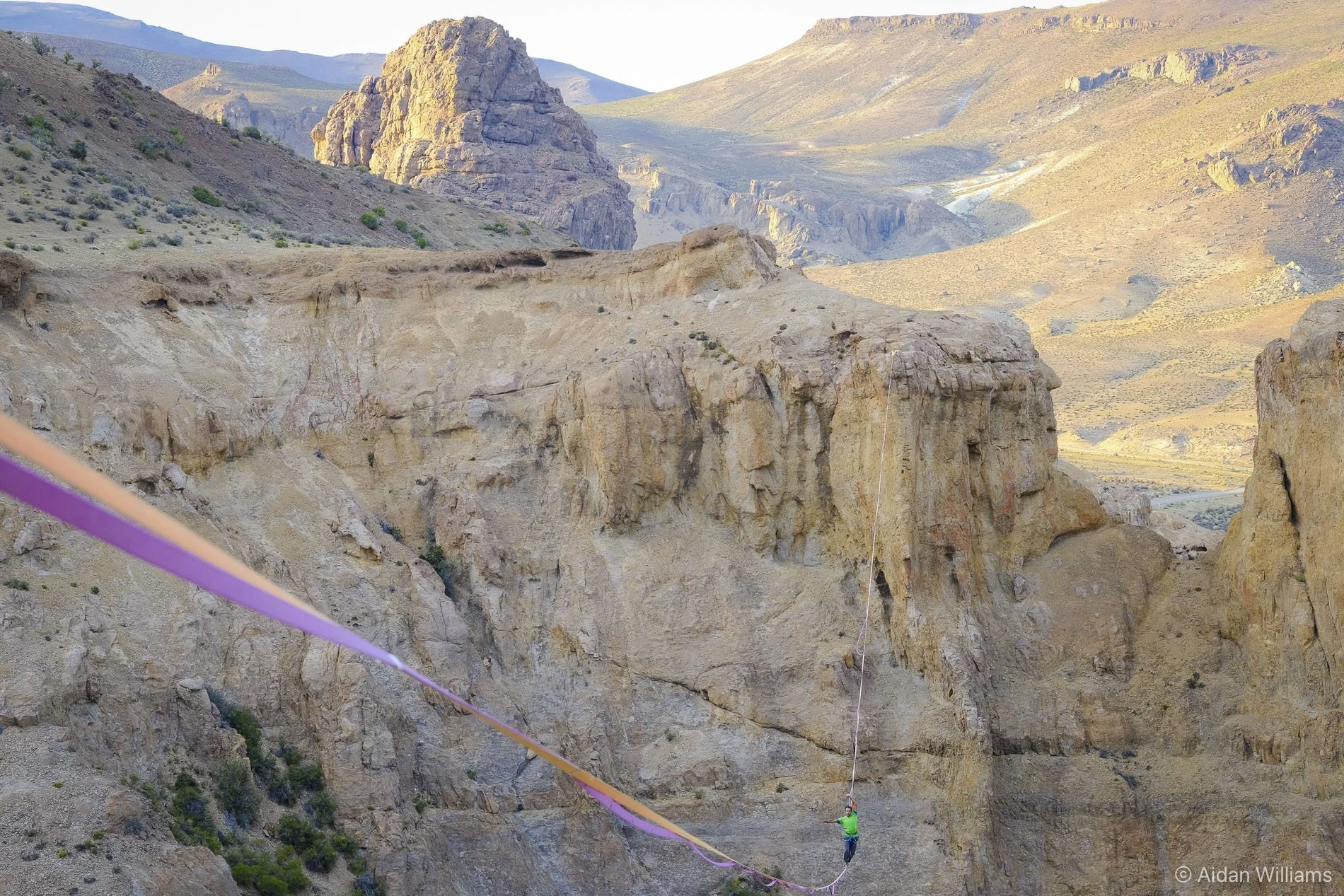 A person in a green shirt bungee jumping from a bridge over a deep canyon with rugged, rocky cliffs and mountains in the background.