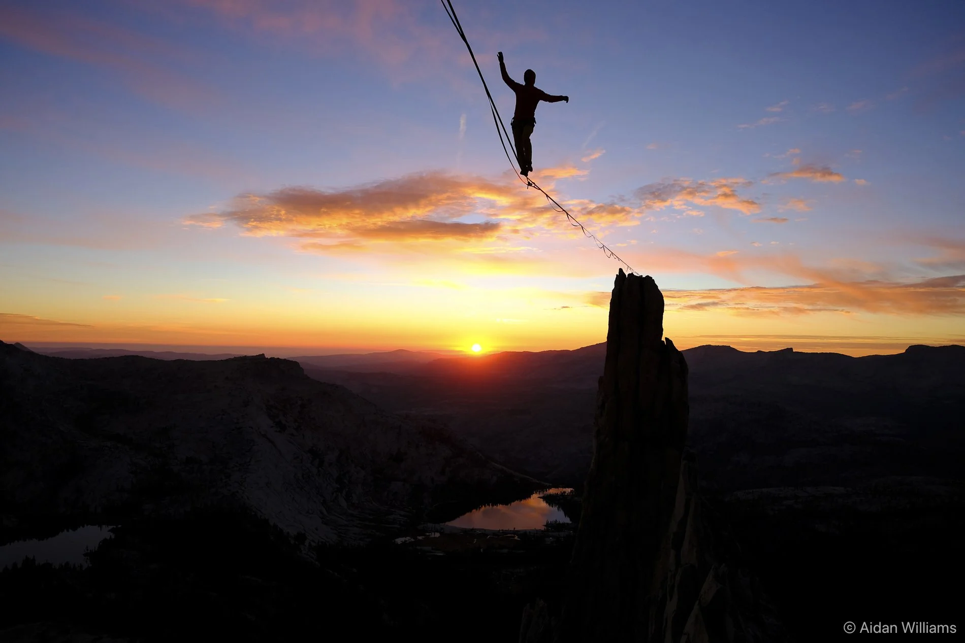 A silhouette of a person walking on a tightrope stretched between a rocky cliff and a high point at sunset or sunrise, with a colorful sky and mountains in the background.