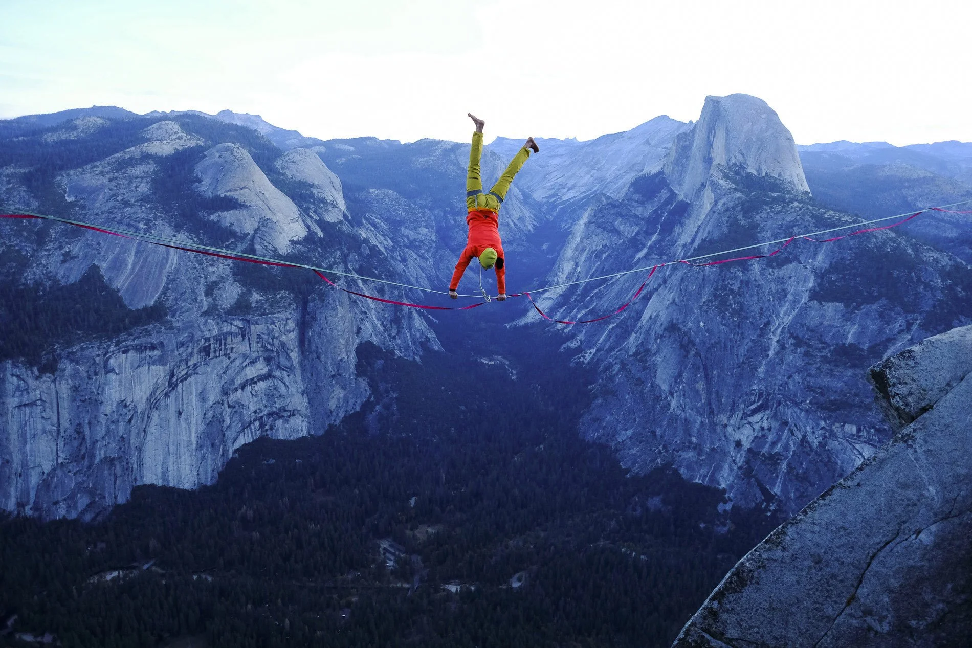 A person in climbing gear balancing upside down on a highline stretched between two mountain cliffs in a mountainous landscape.