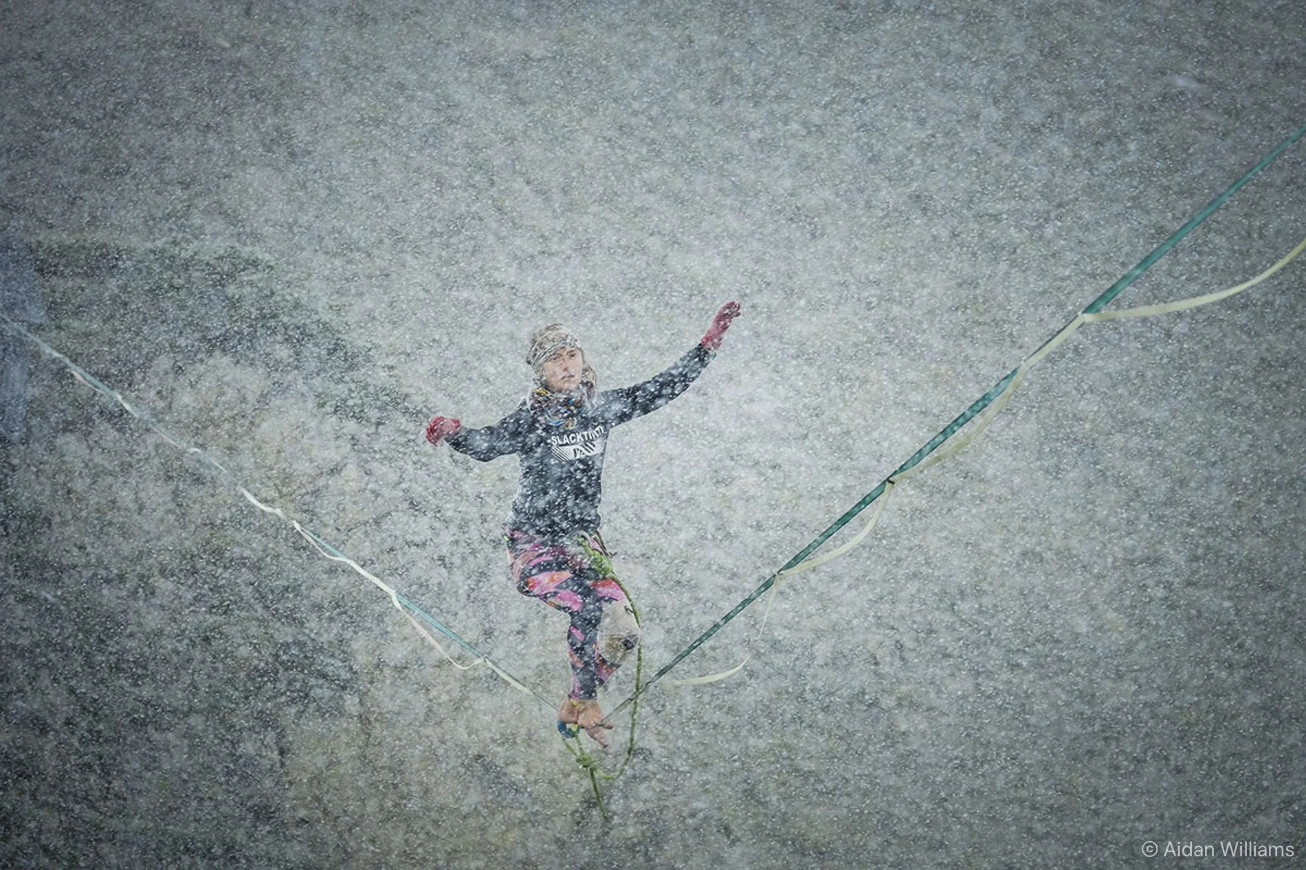A person in athletic clothing and gloves balancing on a slackline in heavy rain.
