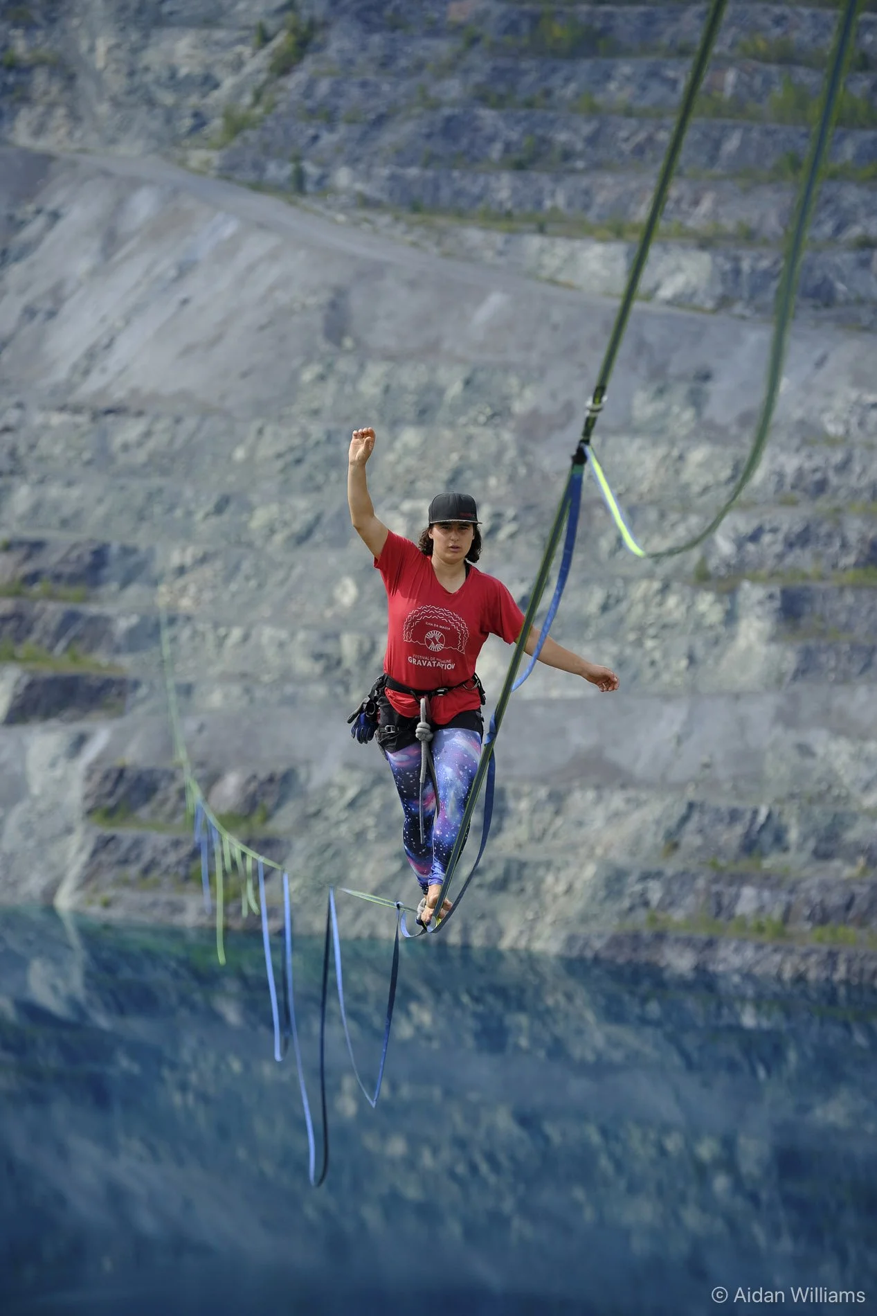 A woman walking on a tightrope above water, wearing starry leggings and holding one arm up, with rocky cliffs in the background.