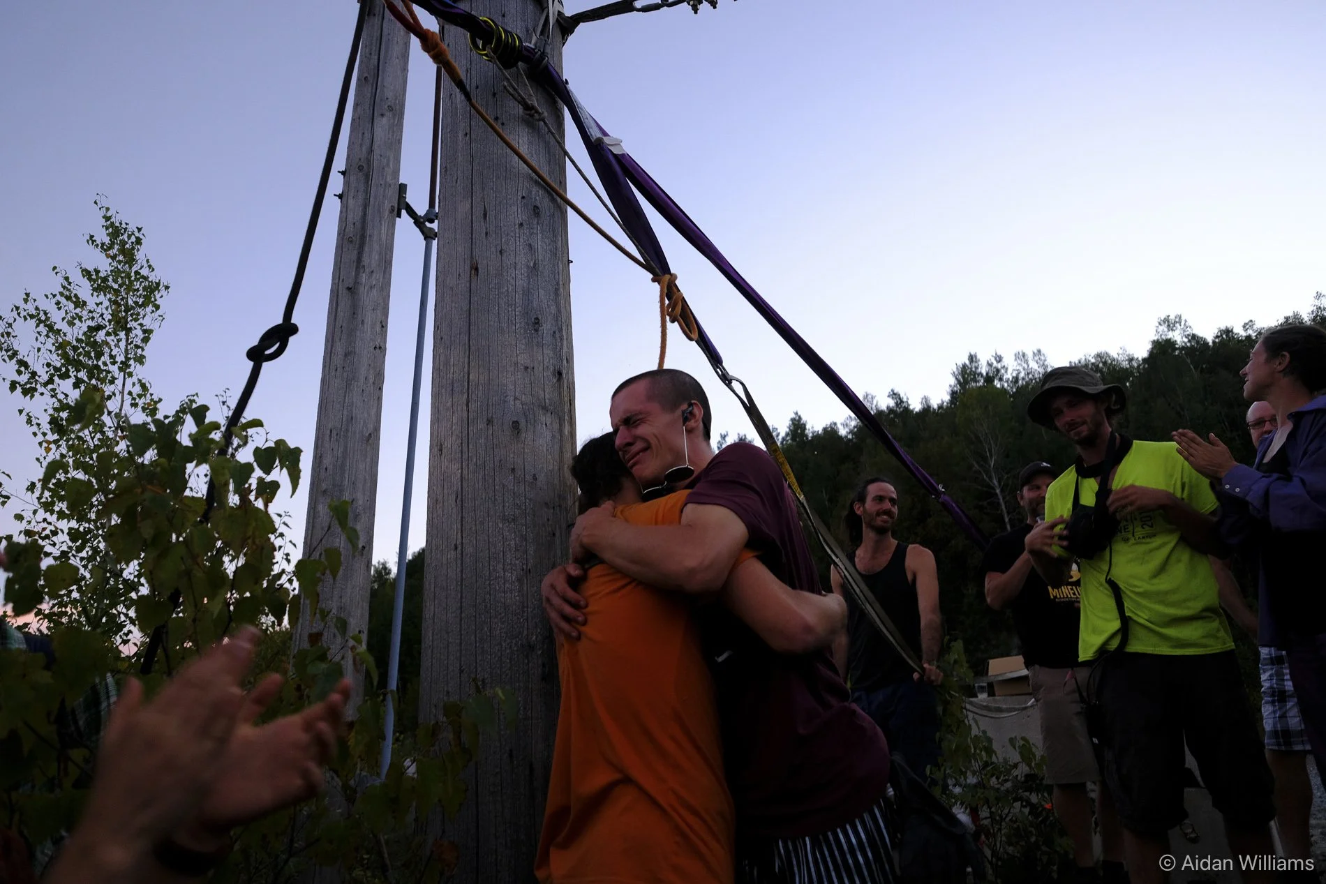 Two people hugging each other in celebration near a wooden utility pole, with several onlookers clapping and smiling around them during dusk.