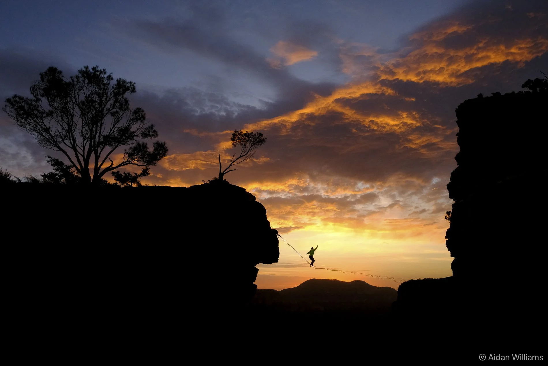 Silhouette of a person balancing on a slackline between two cliffs during sunset, with a colorful sky and trees in the background.
