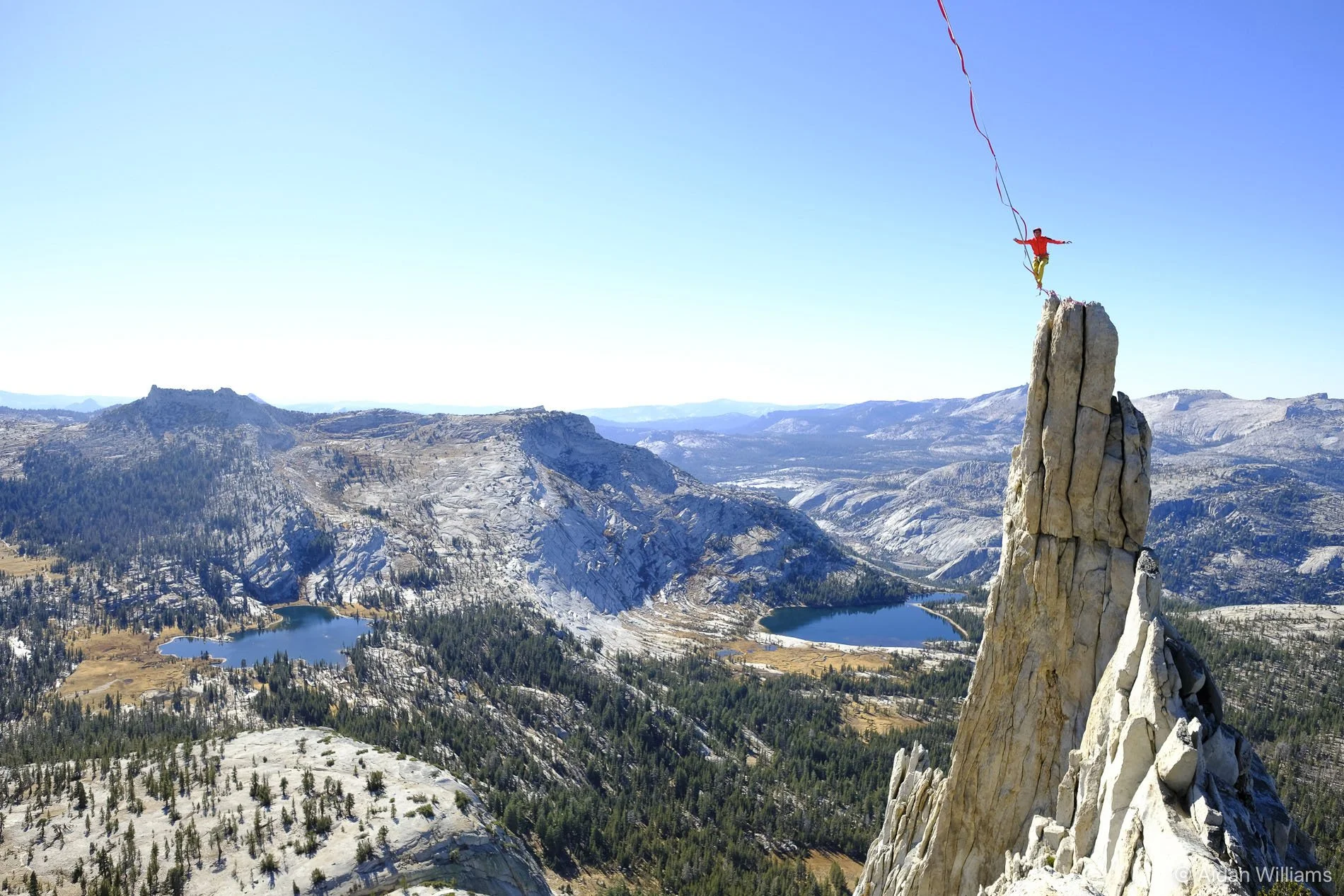 A person in a red jacket and yellow pants standing on a high rock formation, holding a long red ribbon, overlooking a mountainous landscape with lakes and forested areas under a clear blue sky.