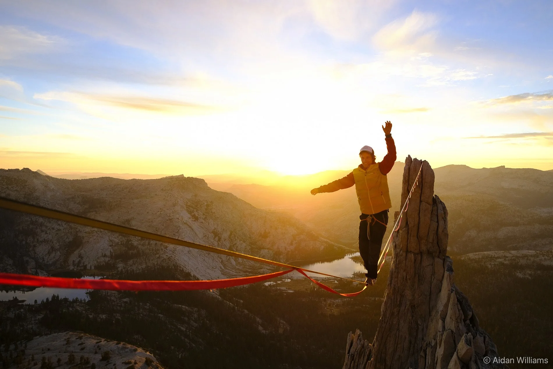 Person balancing with arms extended on a tightrope anchored between a rocky summit during sunset, with scenic mountains and a lake in the background.