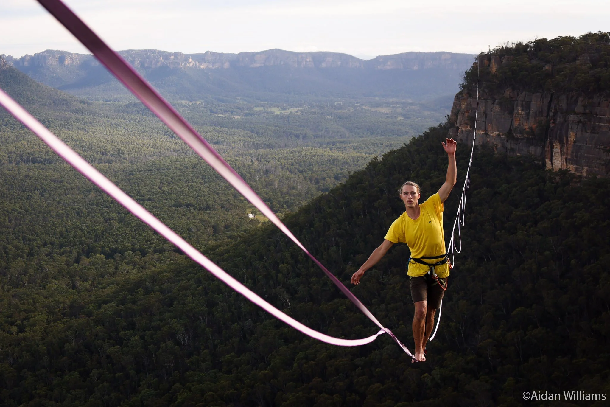 A man walking barefoot on a tightrope stretched high above a lush green valley with distant mountains in the background, surrounded by nature.