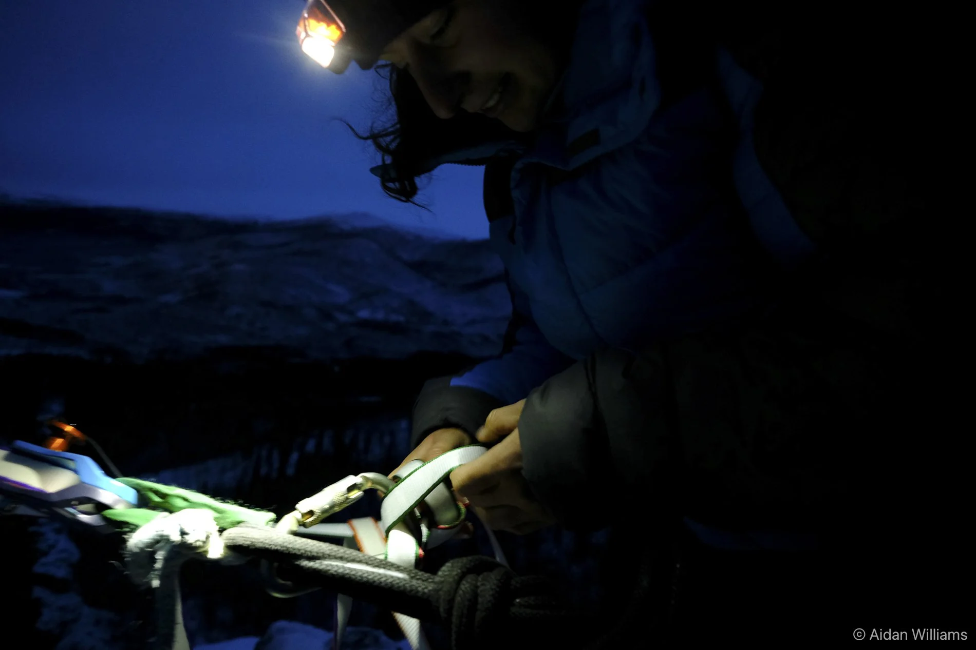 Climber wearing a headlamp adjusting gear on a dark mountain slope at night.
