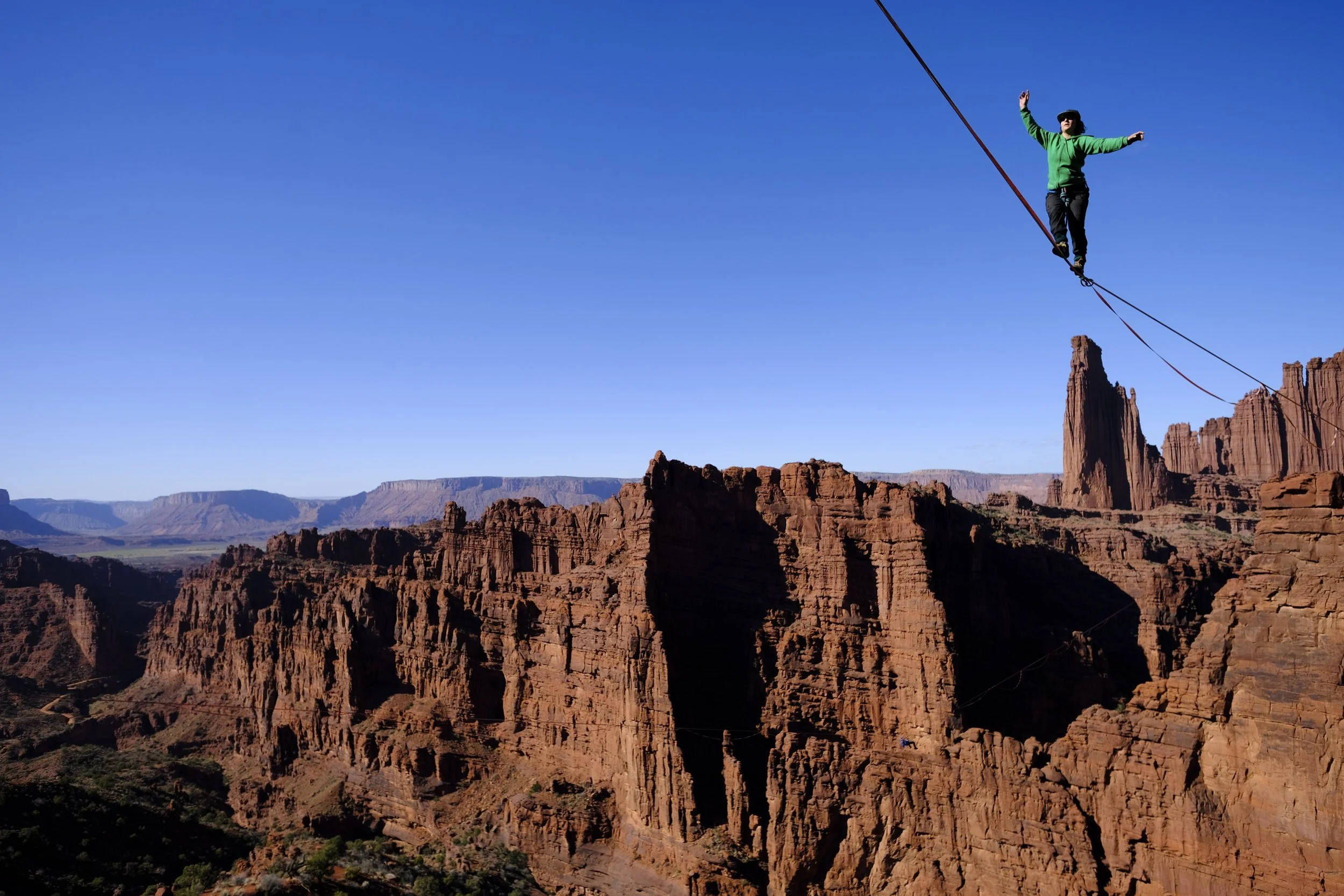 A person walking on a tightrope stretched across red rock canyon formations, with a clear blue sky overhead.