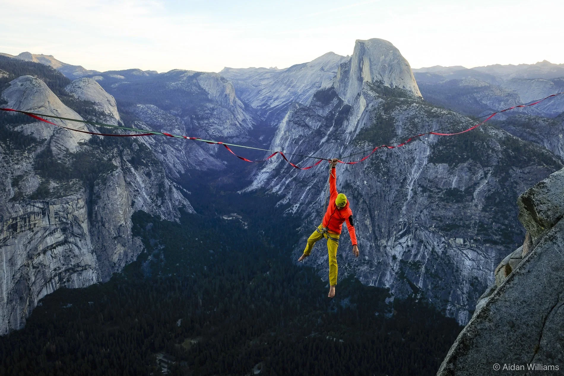 A person dressed in a red jacket and yellow pants, wearing a helmet, is slacklining between two high points in a mountainous landscape, with steep cliff faces and forested valleys in the background.