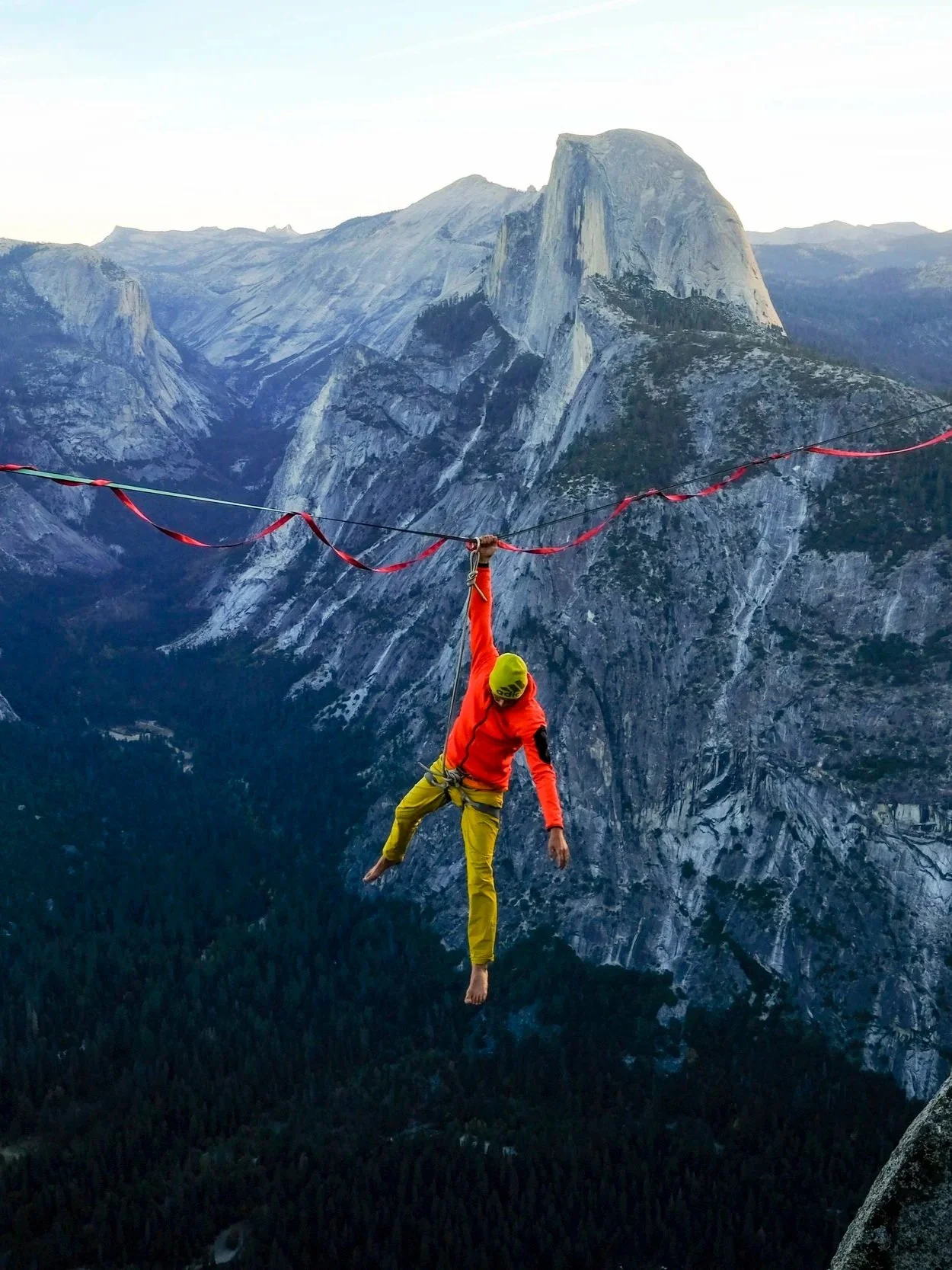 A person in bright outdoor clothing hanging from a high rope between mountain cliffs with a scenic mountain landscape in the background.