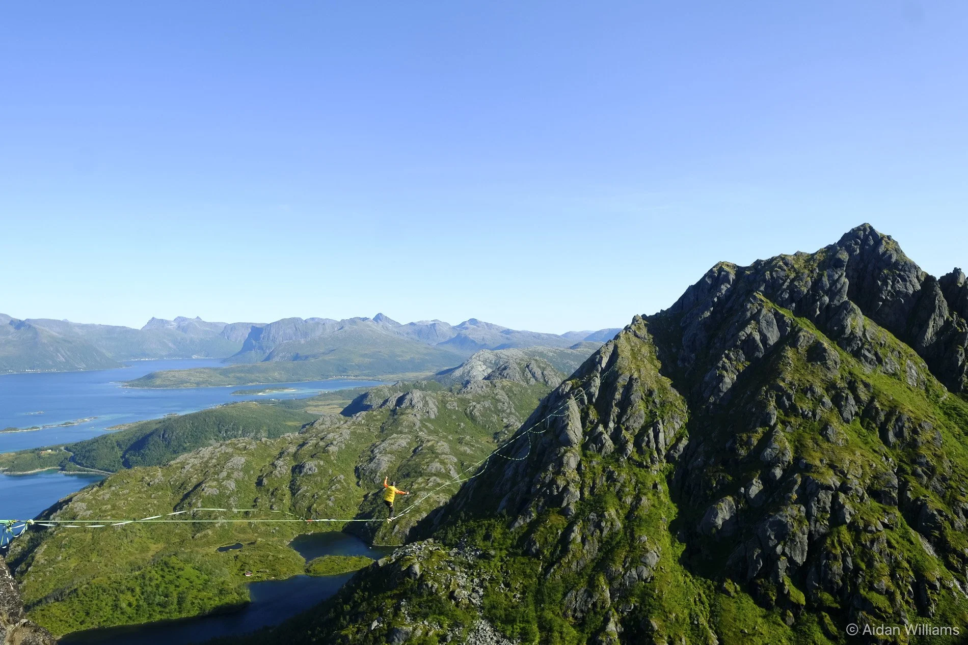 A person walking on a narrow highline between steep green and rocky mountains, with a lake and distant mountains under a clear blue sky.