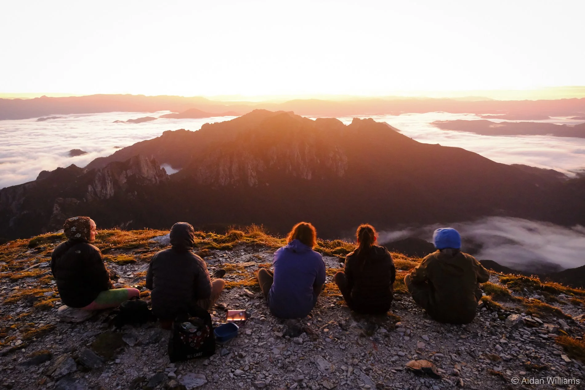 Five people sitting on rocky ground watching sunrise over a mountain range with clouds below.
