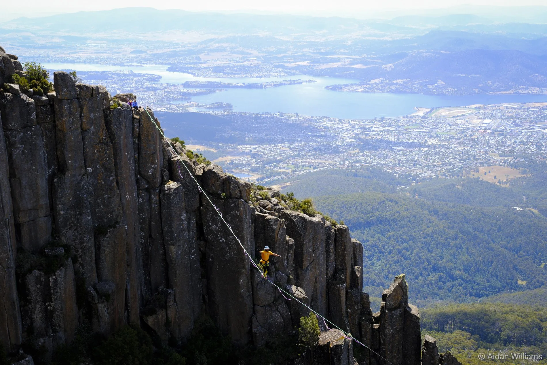 Climber in yellow jacket securing to a rope on a steep rocky mountain face with a scenic view of a lake and city below