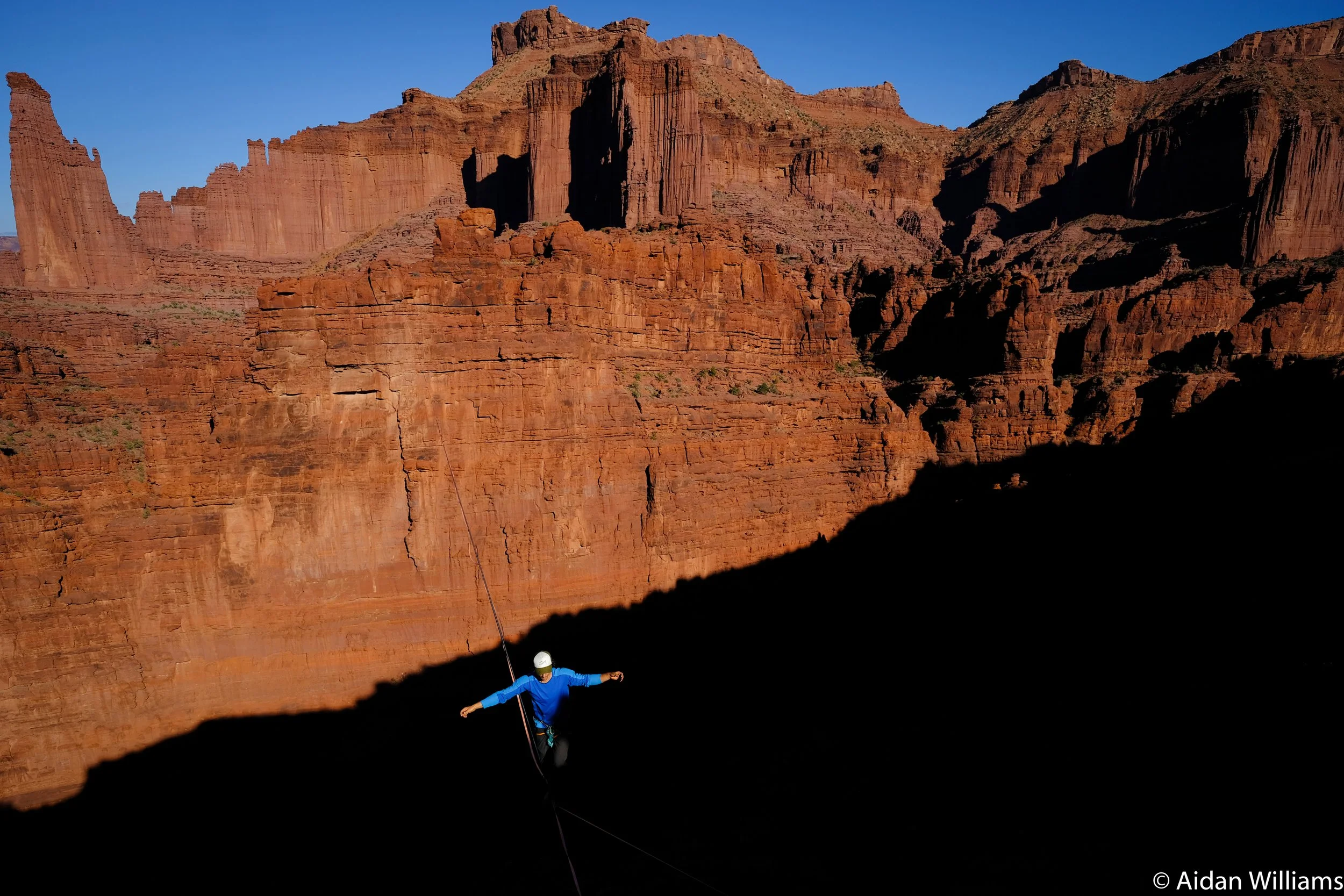 A person in a blue jacket and white helmet is bungee jumping over a deep canyon with red rock cliffs in the background, casting a shadow across part of the rock formation.