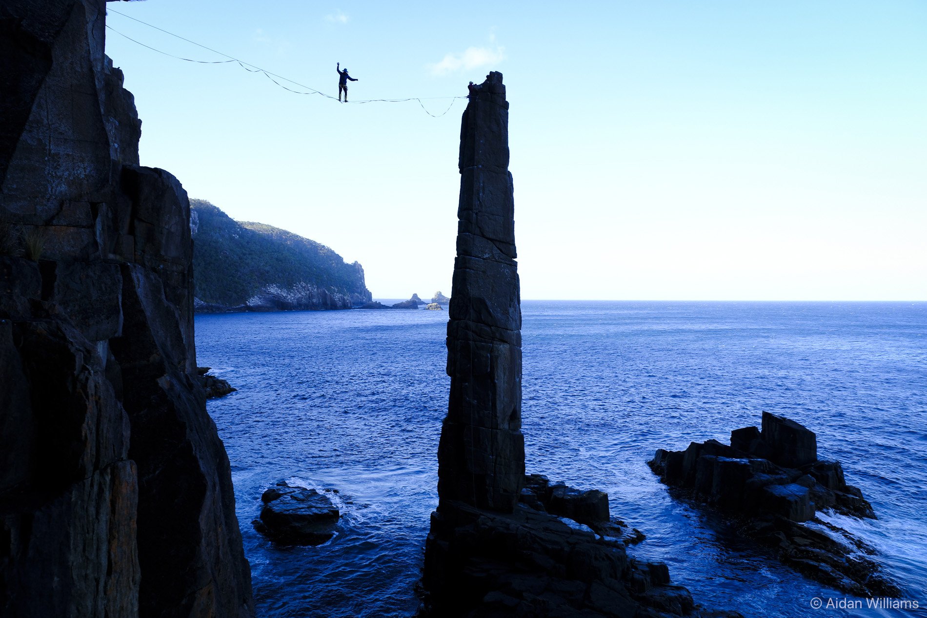 A person balancing on a high tightrope stretched between two large rocky cliffs over the ocean, with one arm raised and the other holding onto the tightrope, and a forested headland in the background.