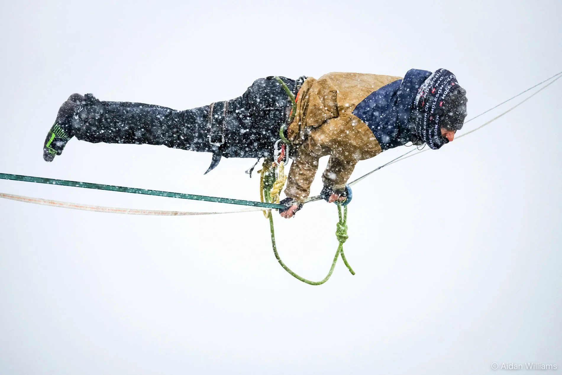 A person wearing winter clothing, including a jacket, gloves, and a beanie, is climbing a snowy mountain with ropes and safety gear in a snowy, foggy environment.