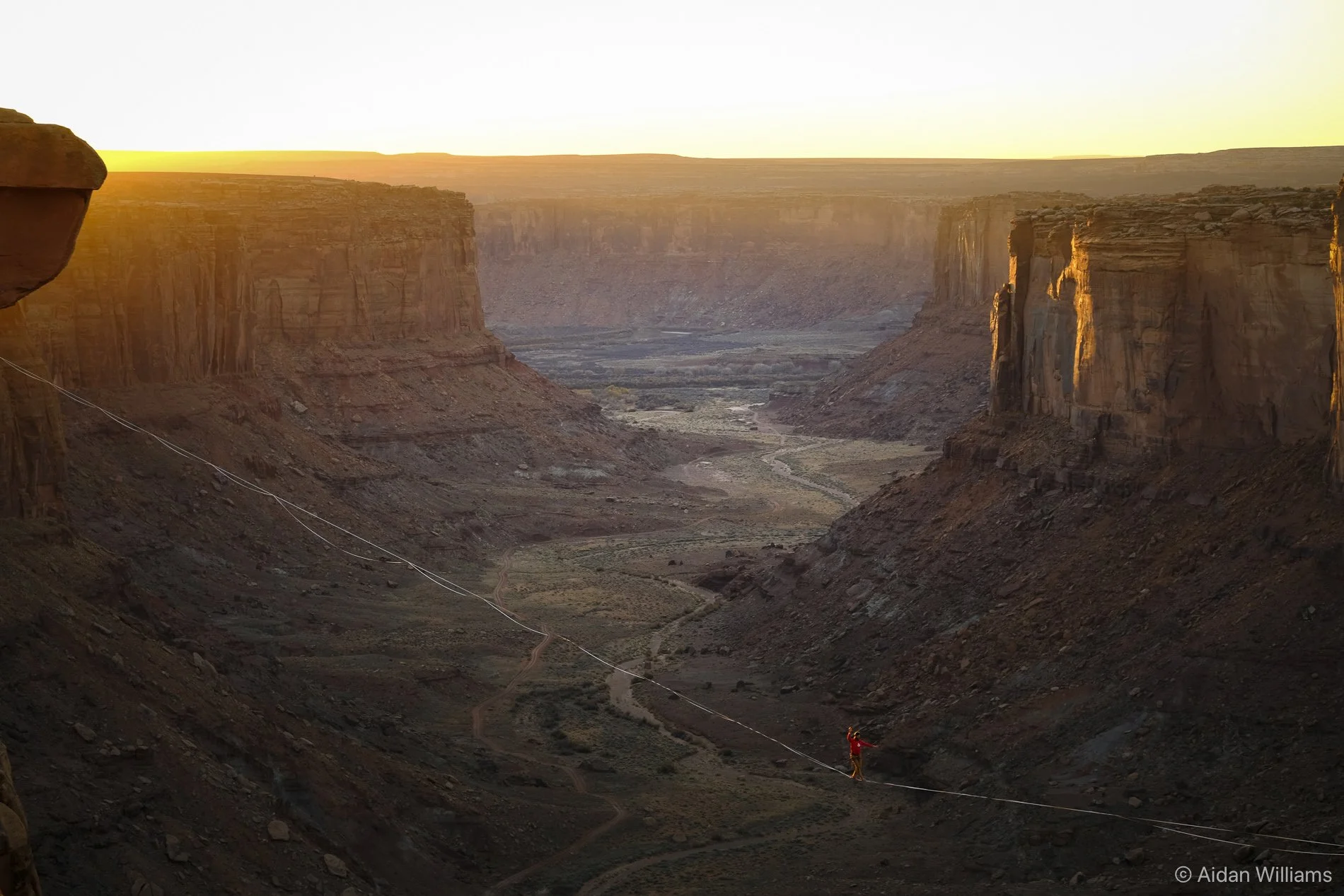 A person crossing a tightrope high above a desert canyon during sunset.