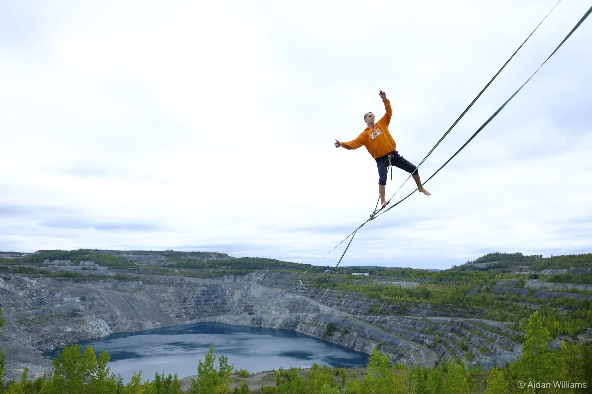 A man walking on a tightrope high above a quarry, with a body of water and quarry walls in the background.
