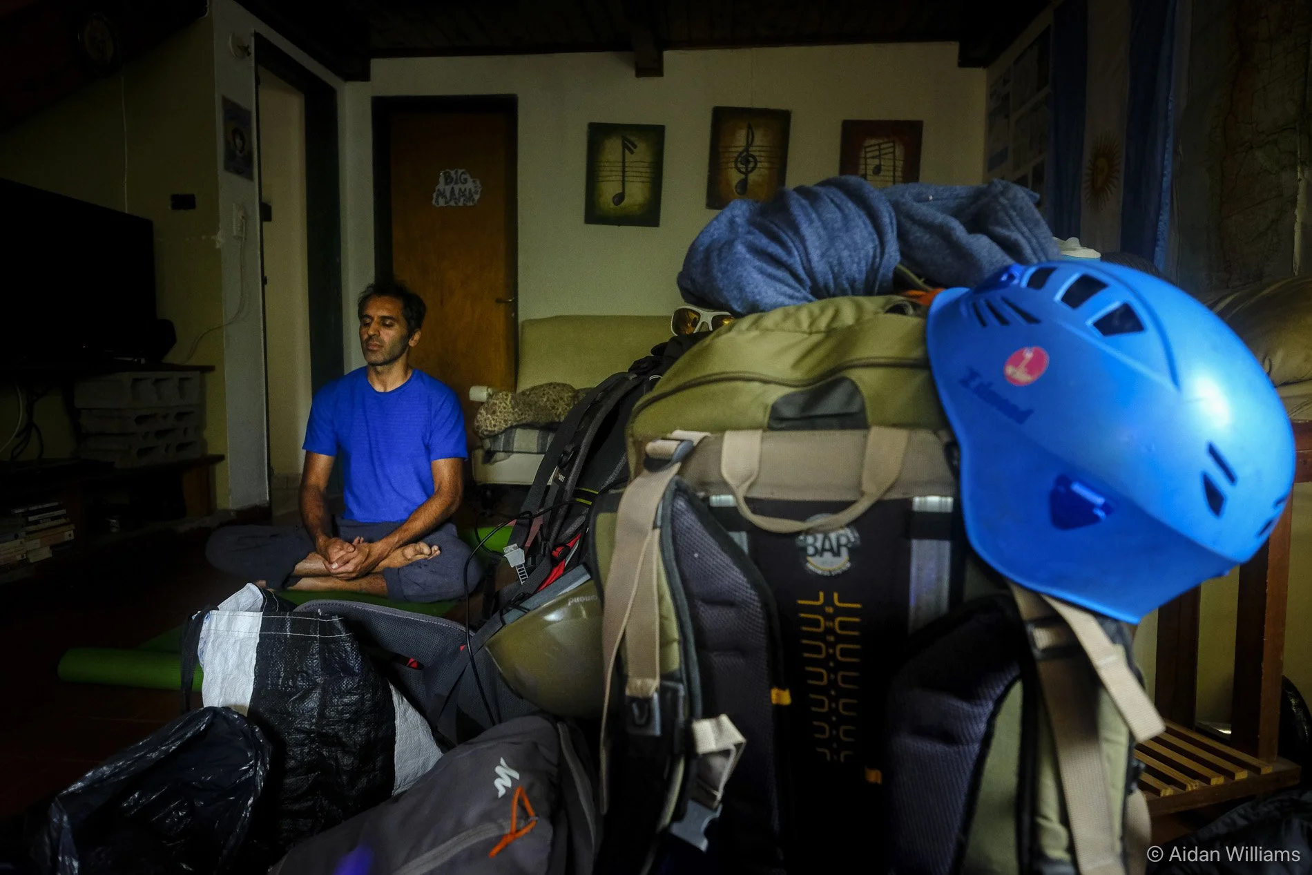 A man wearing a blue shirt meditating in a room with musical notes on the wall behind him. The room has a door labeled 'Big Mama' and is filled with packed backpacks, helmets, and outdoor gear.