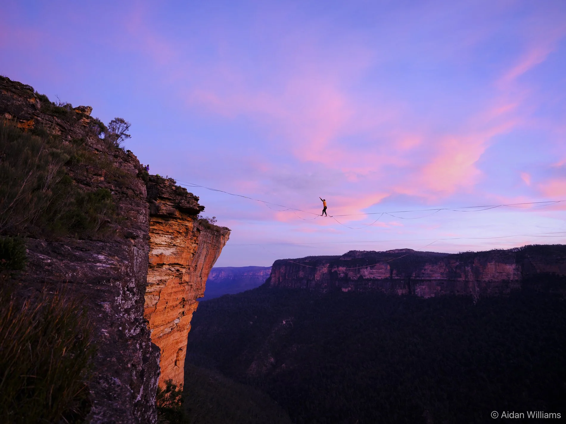 A person walking on a tightrope stretched across a deep canyon during sunset, with cliffs and colorful sky in the background.
