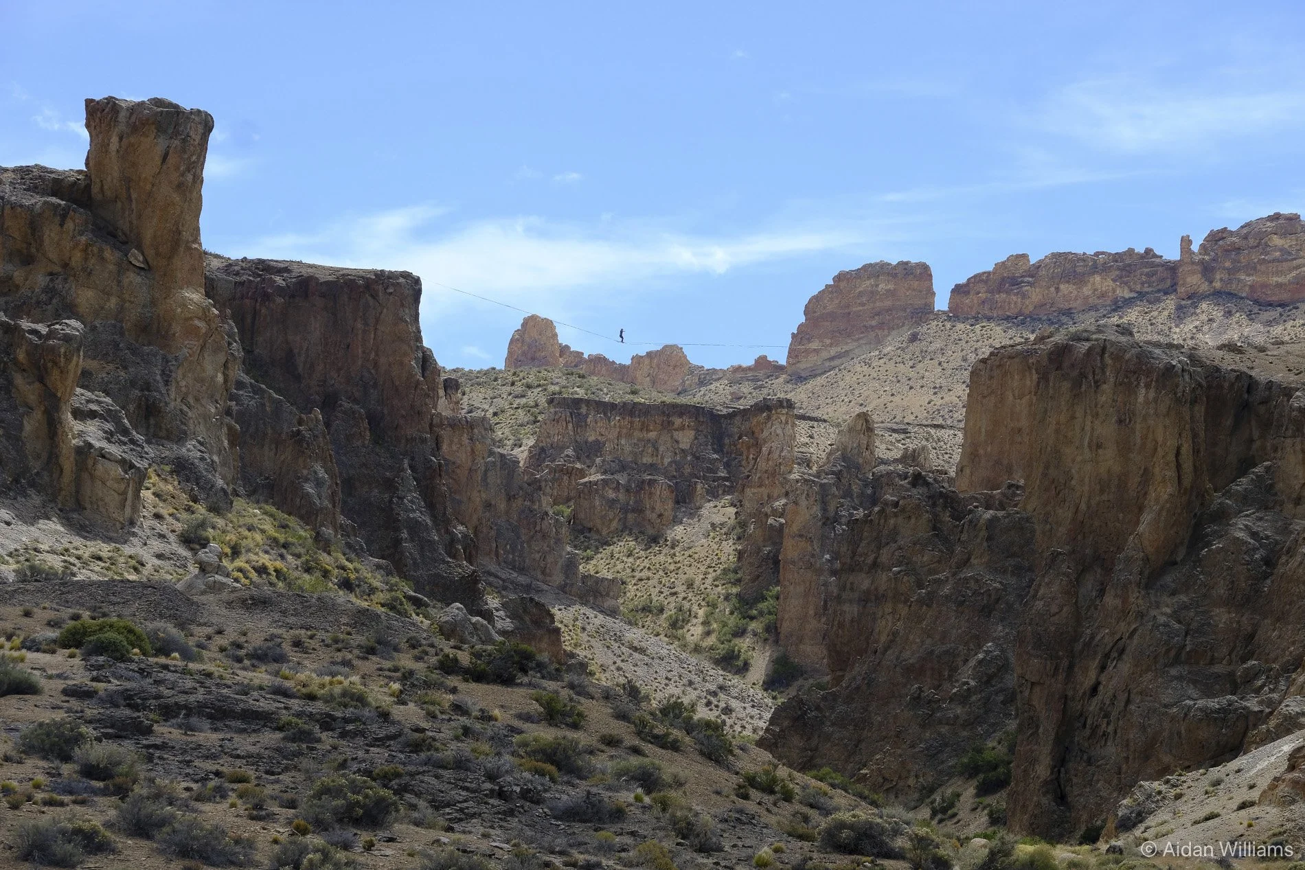 A person walking on a tightrope stretched across a canyon with rocky cliffs and sparse desert vegetation under a blue sky.