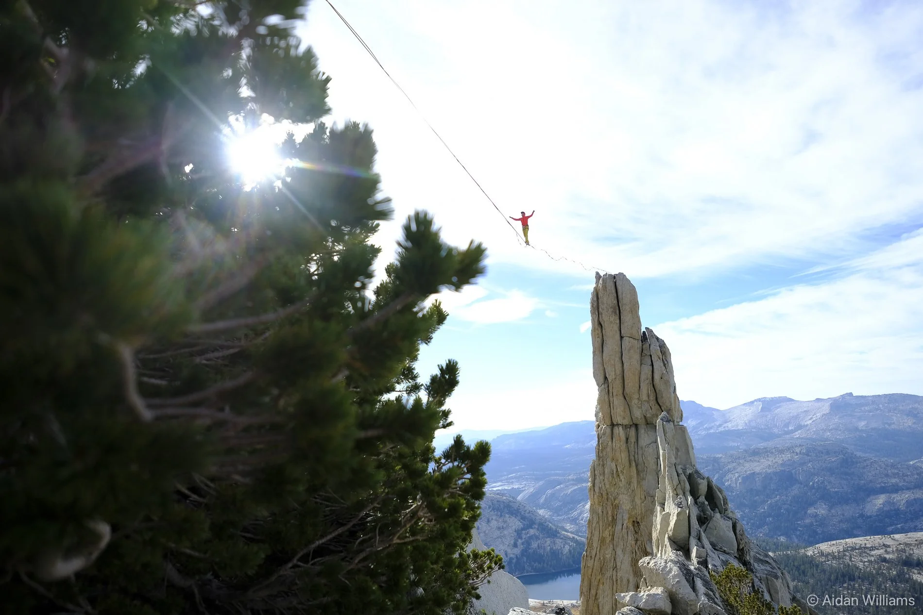 A person walking on a tightrope stretched between a rocky peak and a tree with the sun shining through the branches, in a mountain landscape with blue sky and distant mountains.