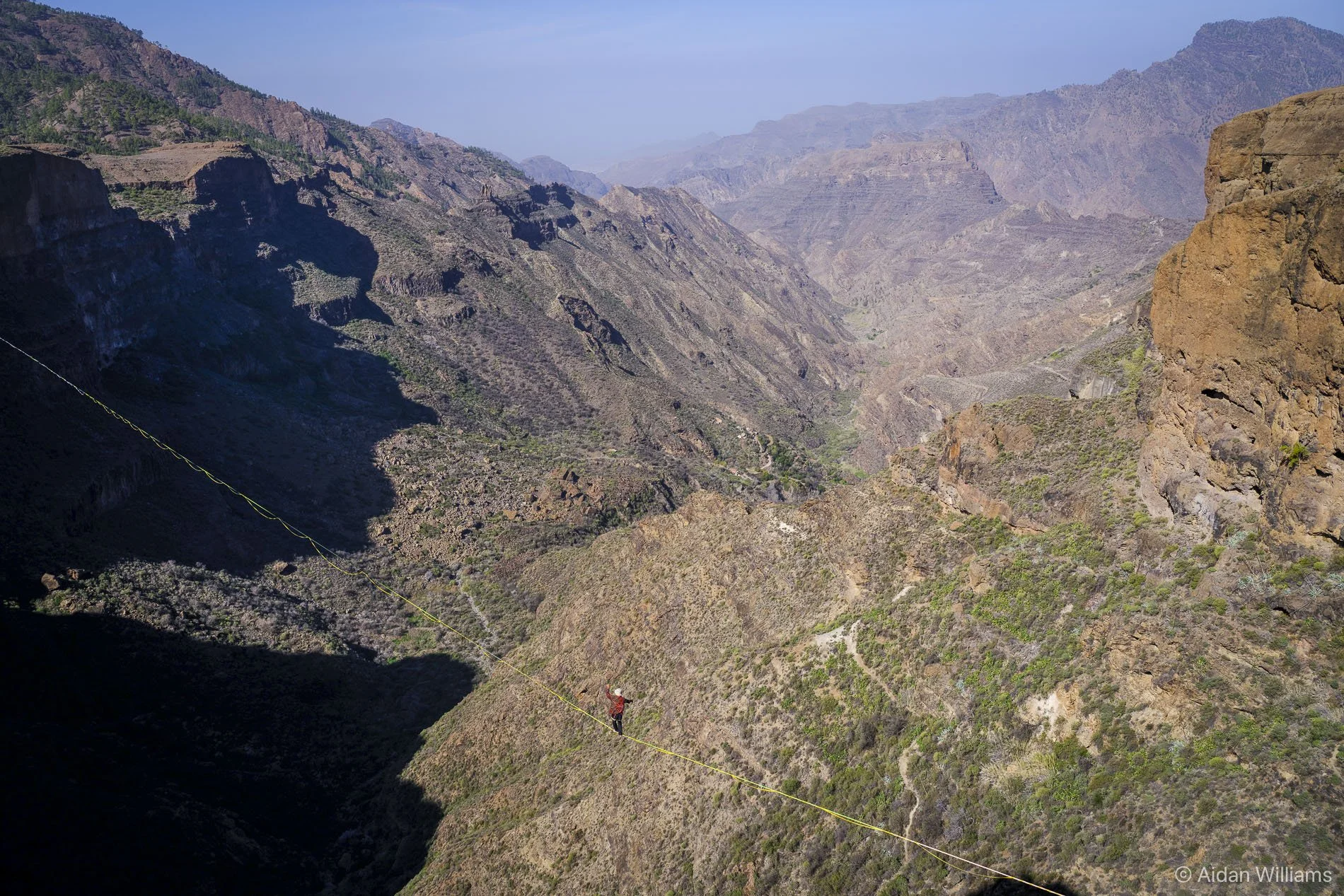 A person walking on a narrow cable bridge high above a rugged canyon with cliffs and mountainous terrain.