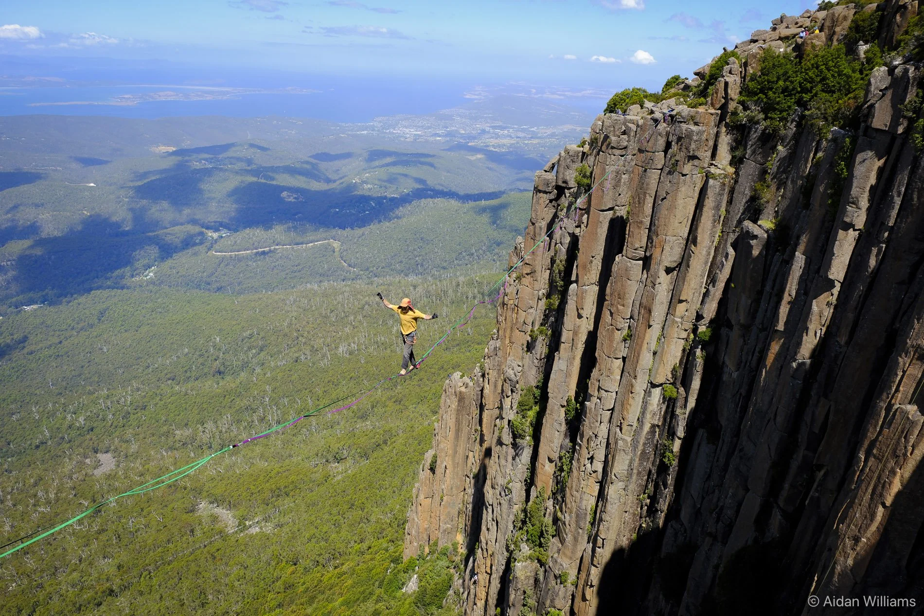 A person wearing a yellow shirt and hat walking on a slackline stretched between a tall rocky cliff and another point in the distance, high above a green forested landscape with a view of water and distant land in the background.