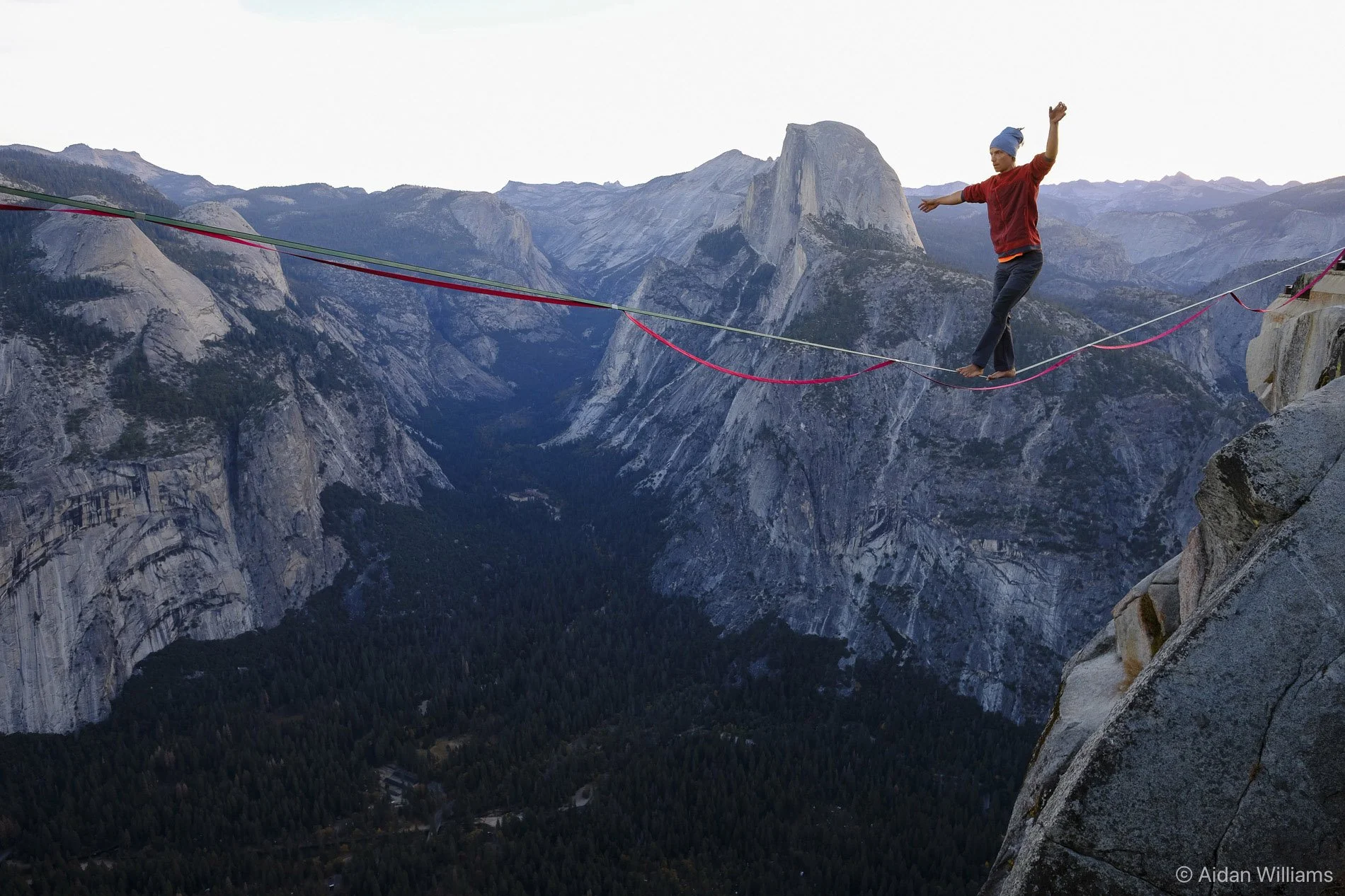 Person walking on a slackline stretched between two mountain cliffs with a mountainous landscape in the background.