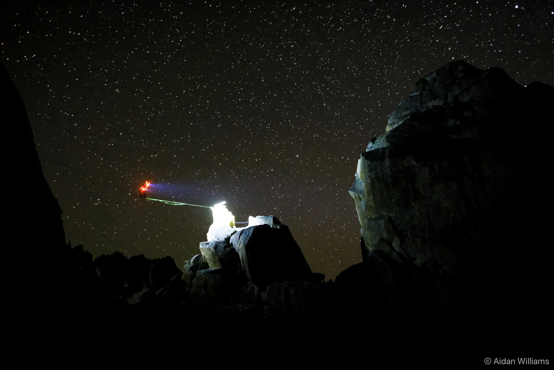 Night sky filled with stars above rugged mountain terrain with climbers or explorers setting up a camp or climbing equipment, illuminated by artificial light.