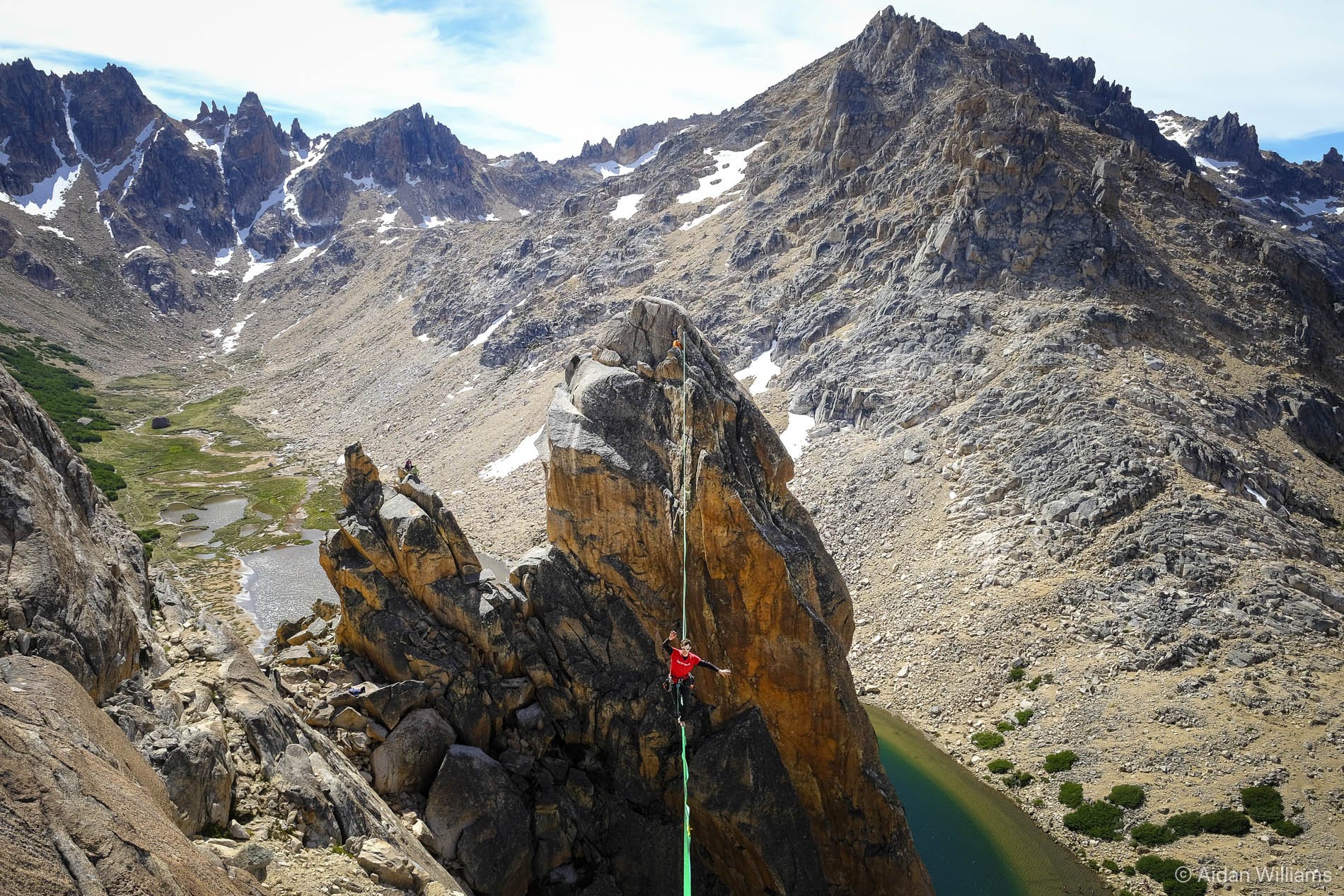 A climber in a red jacket and helmet ascending a steep rock face with a rope in a mountain landscape with rugged peaks, patches of snow, a green valley, and a lake.