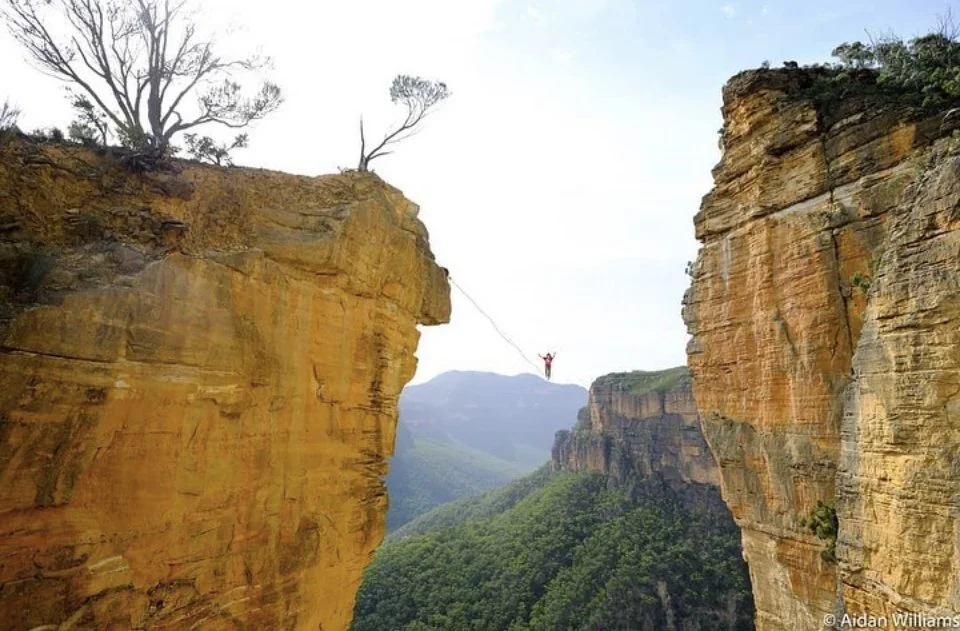 A person on a zip line high above a deep canyon with steep, rocky cliffs and green forested valley below.