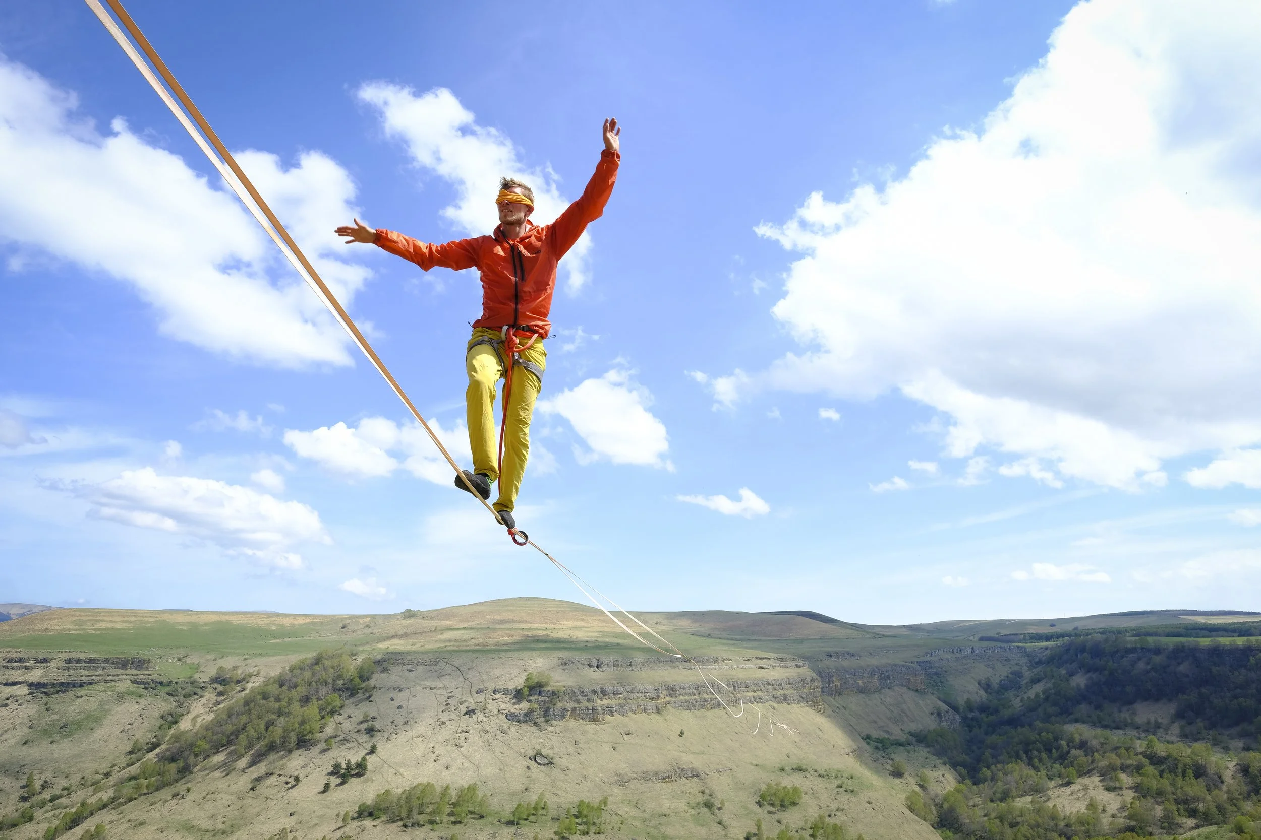 Person walking on a tightrope high above a landscape of hills and valleys, wearing colorful outdoor gear and a blindfold.