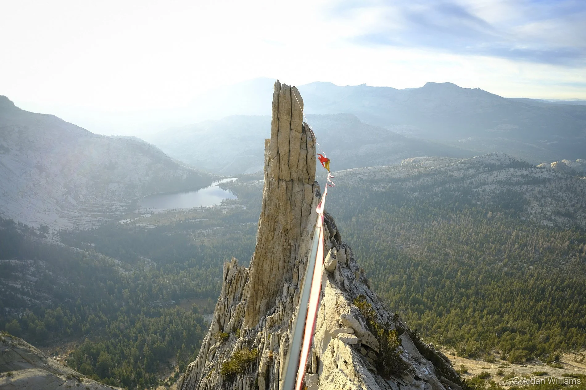 View from a mountain summit with a rocky, steep edge in the foreground; a narrow safety cable runs along the edge; forested mountains and a lake are in the distance under a partly cloudy sky.