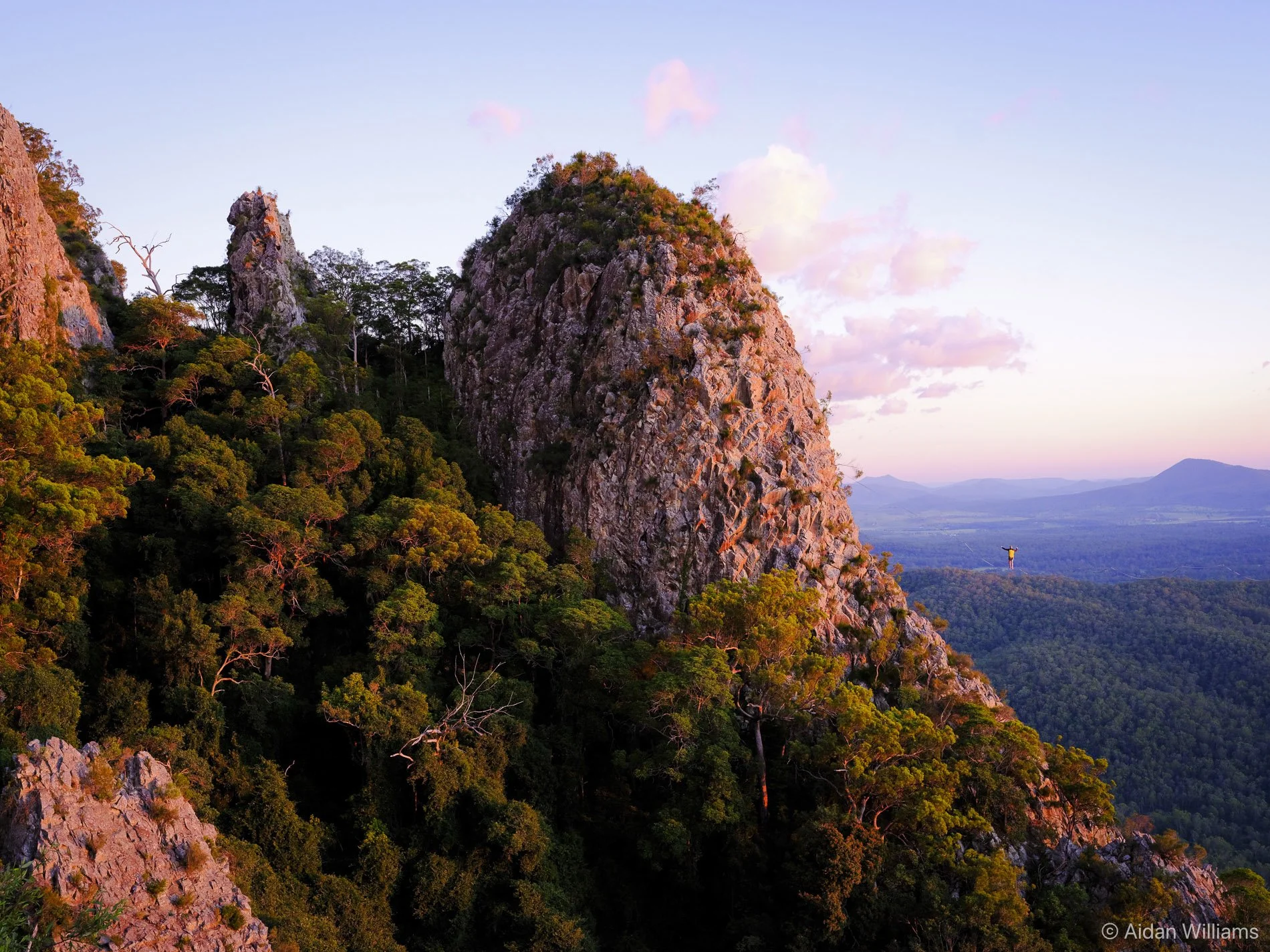 Sunset over rugged mountain landscape with dense green forest and sky with pink clouds.
