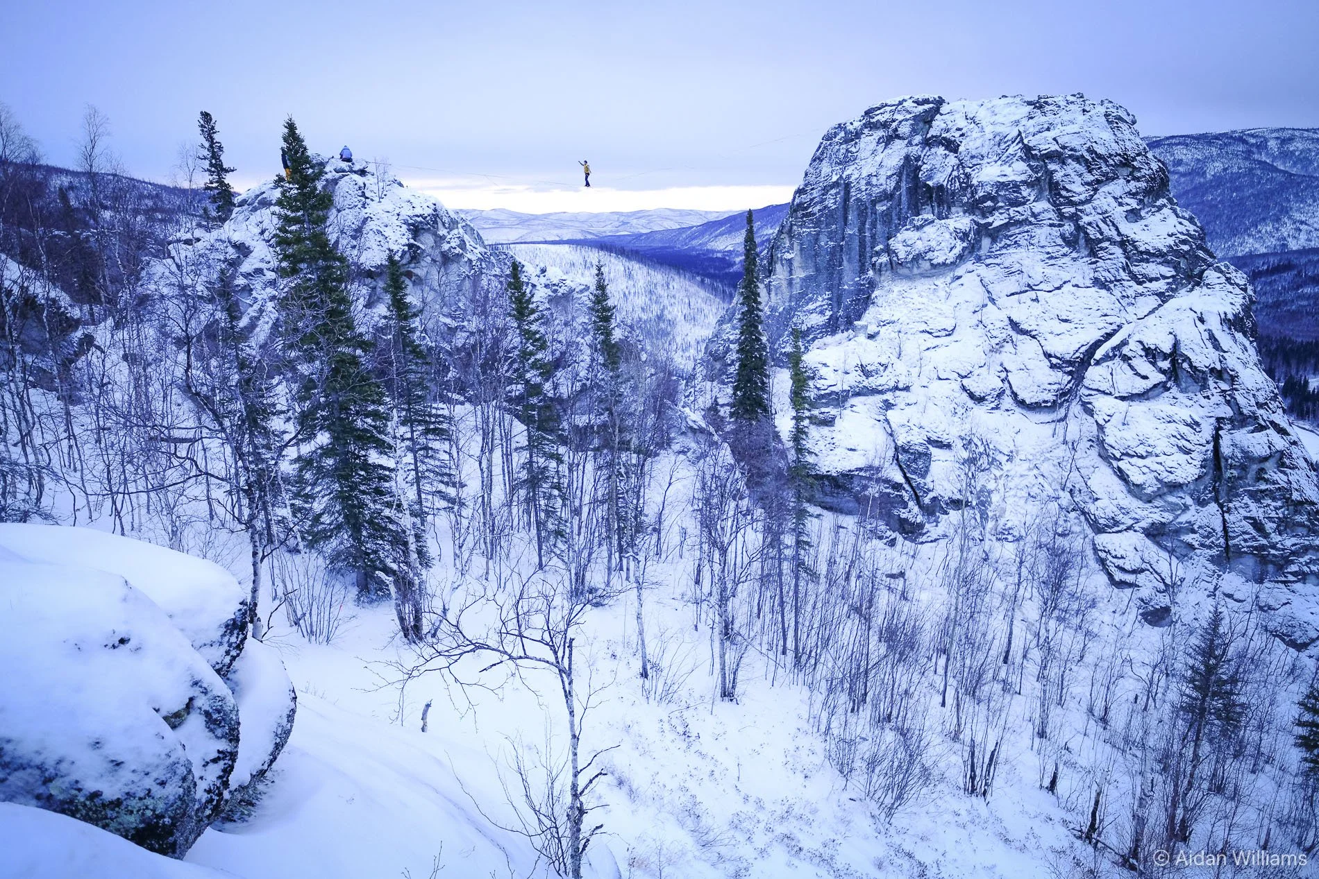 Snow-covered mountainous landscape with rocky cliffs, pine trees, and two people on a zip line in the distance.