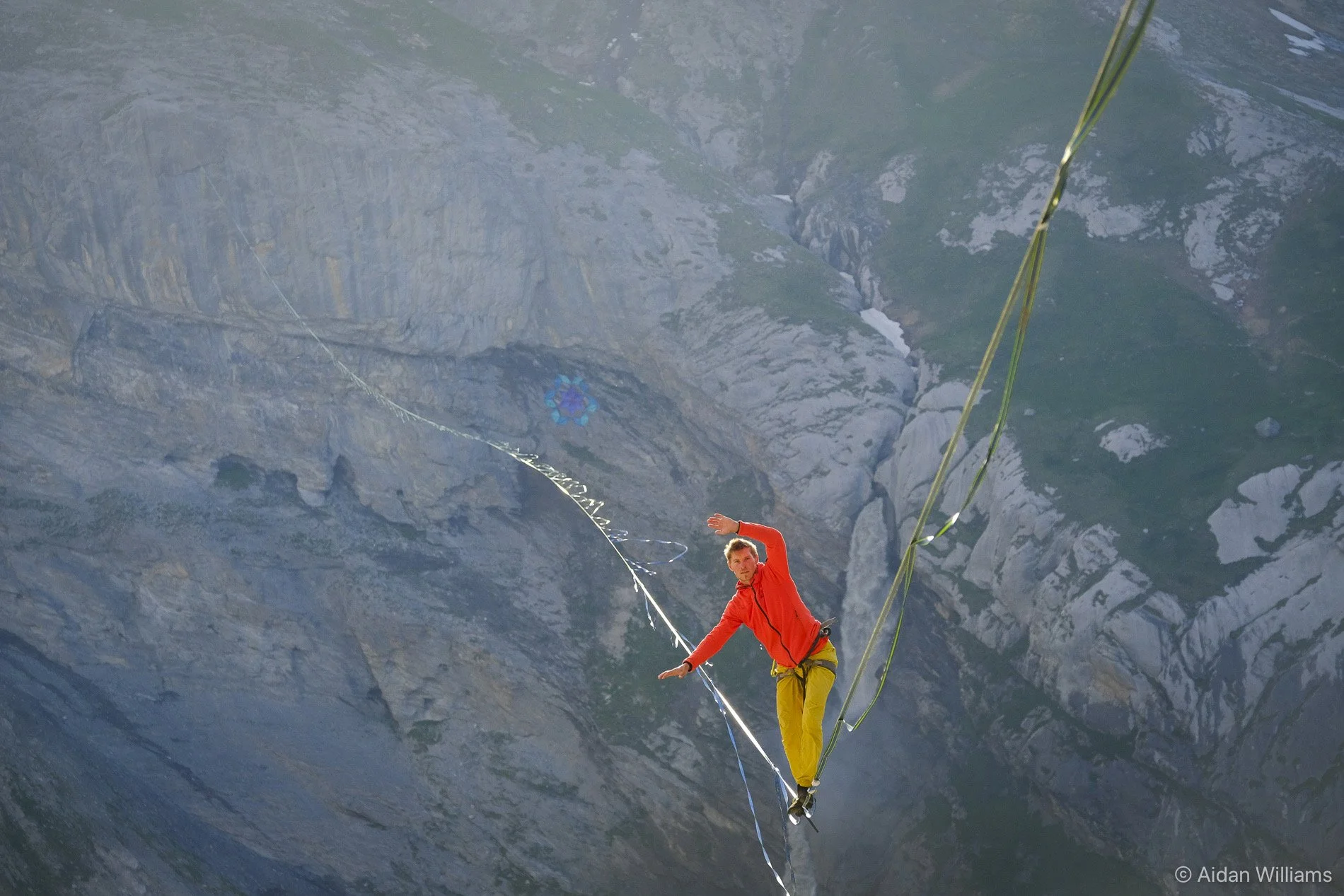 A person in a red jacket and yellow pants balancing on a tightrope high above a mountain landscape with rocky cliffs and steep slopes.