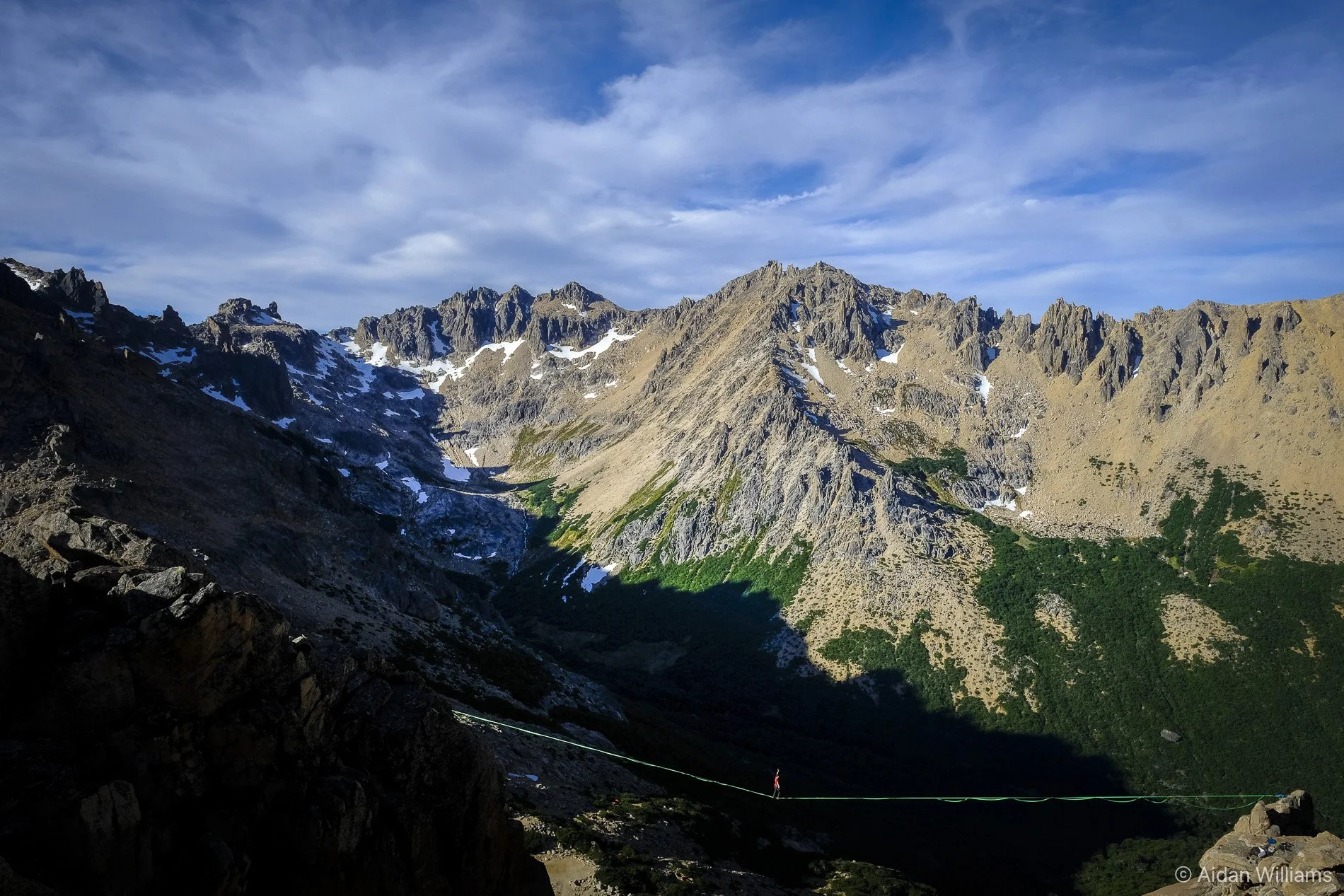 A rugged mountain landscape with steep rocky peaks, patches of snow, green vegetation on the lower slopes, and a partly cloudy sky overhead.