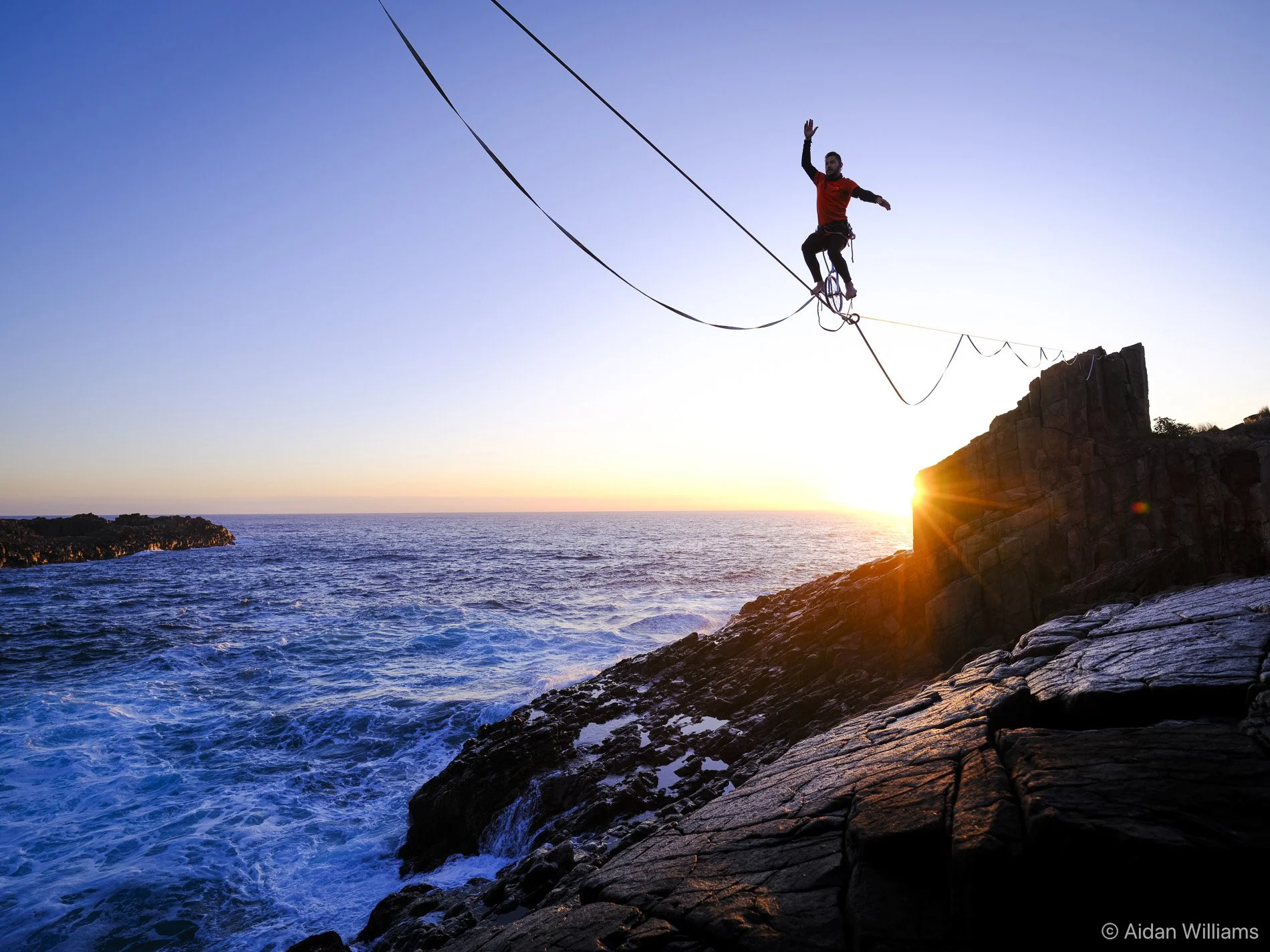 A person walking on a slackline over the ocean at sunset, balancing on a thin rope attached to rocks, with the sun setting on the horizon.