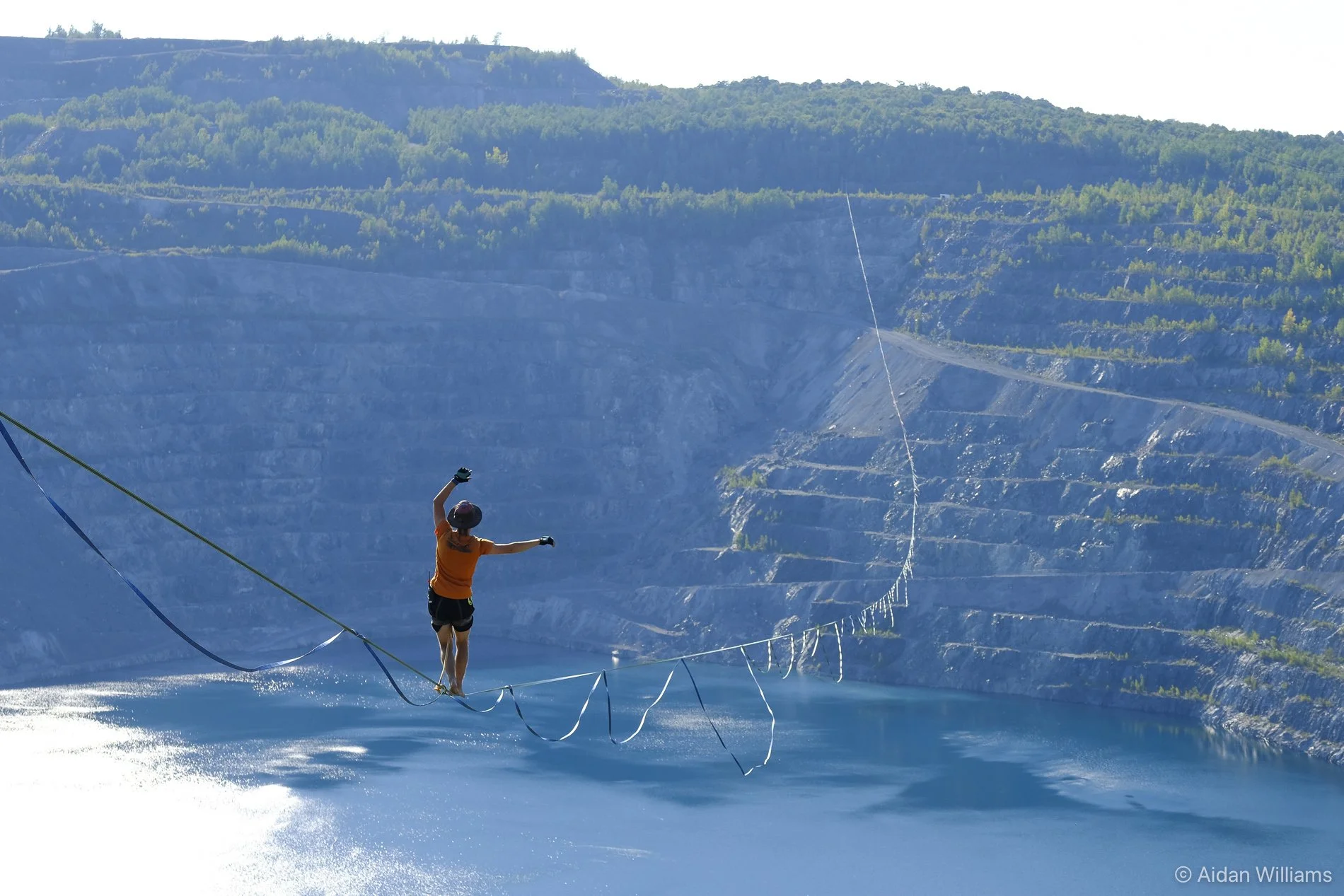 A person wearing a helmet and orange shirt tightrope walking over water with a safety harness, a canyon with layered cliffs and trees in the background.