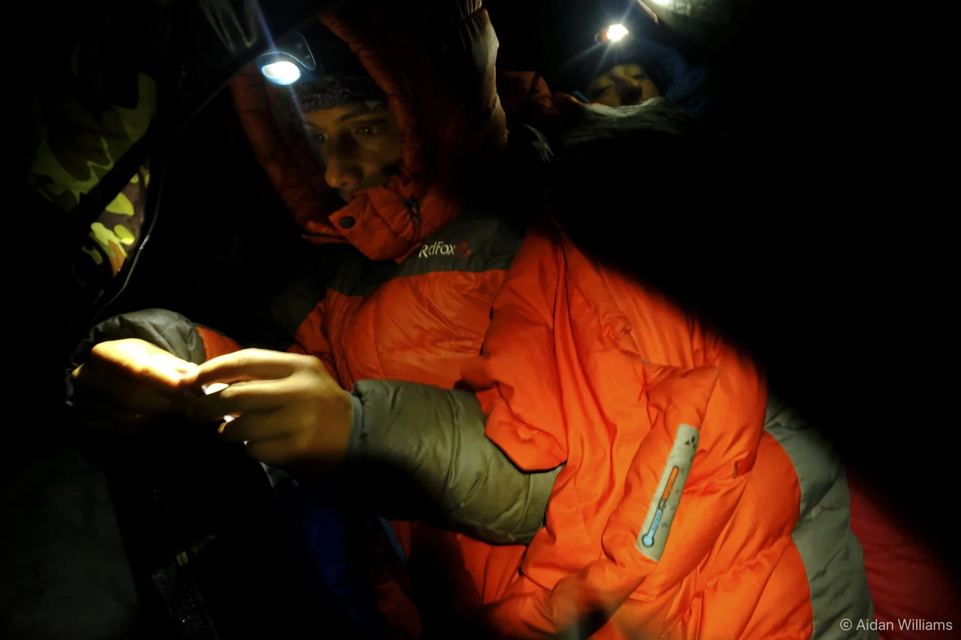 A person wearing a red hooded jacket and sleeping bag with a thermometer patch, lying in a tent with a headlamp, looking at a smartphone.