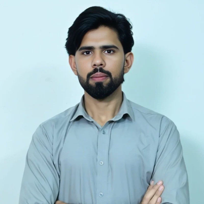 A young man with dark hair and a beard, wearing a light gray button-up shirt, standing with arms crossed in front of a plain light blue background.