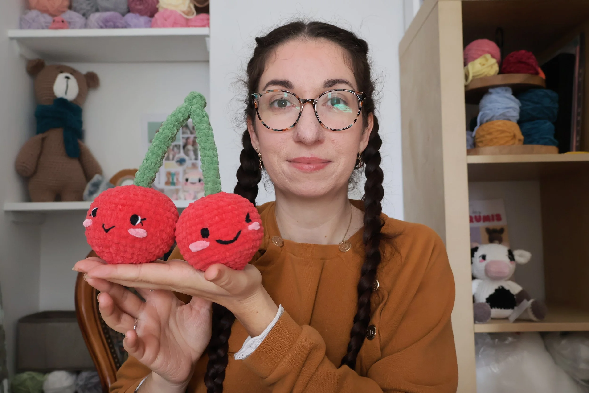 Une femme avec des lunettes et des cheveux tressés, tenant deux peluches de cerises crochetées avec des visages souriants, dans un environnement intérieur avec des étagères remplies de peluches et de pelotes de laine.