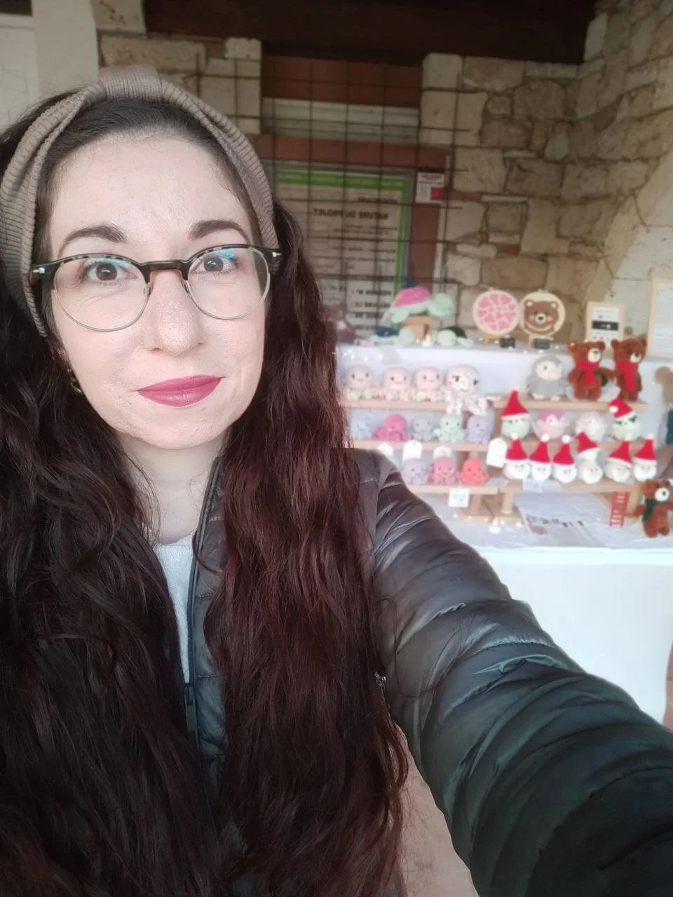 Une femme portant des lunettes, un bandeau et un manteau prend une selfie près d'un stand avec des décorations de Noël et des jouets en peluche de Noël et d'hiver.
