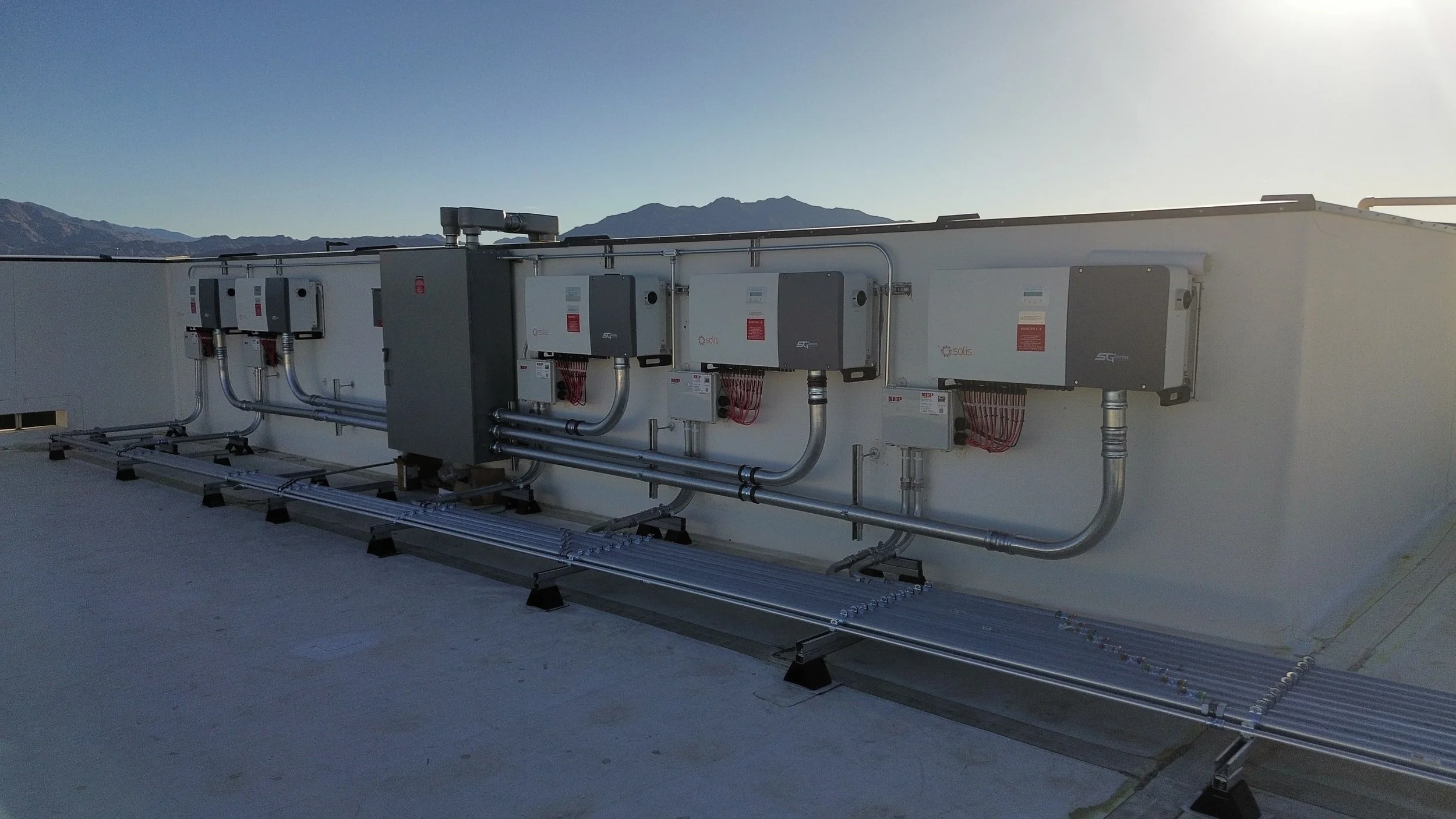 Commercial solar power inverter system on rooftop with electrical conduit and wiring, with mountain landscape in background.