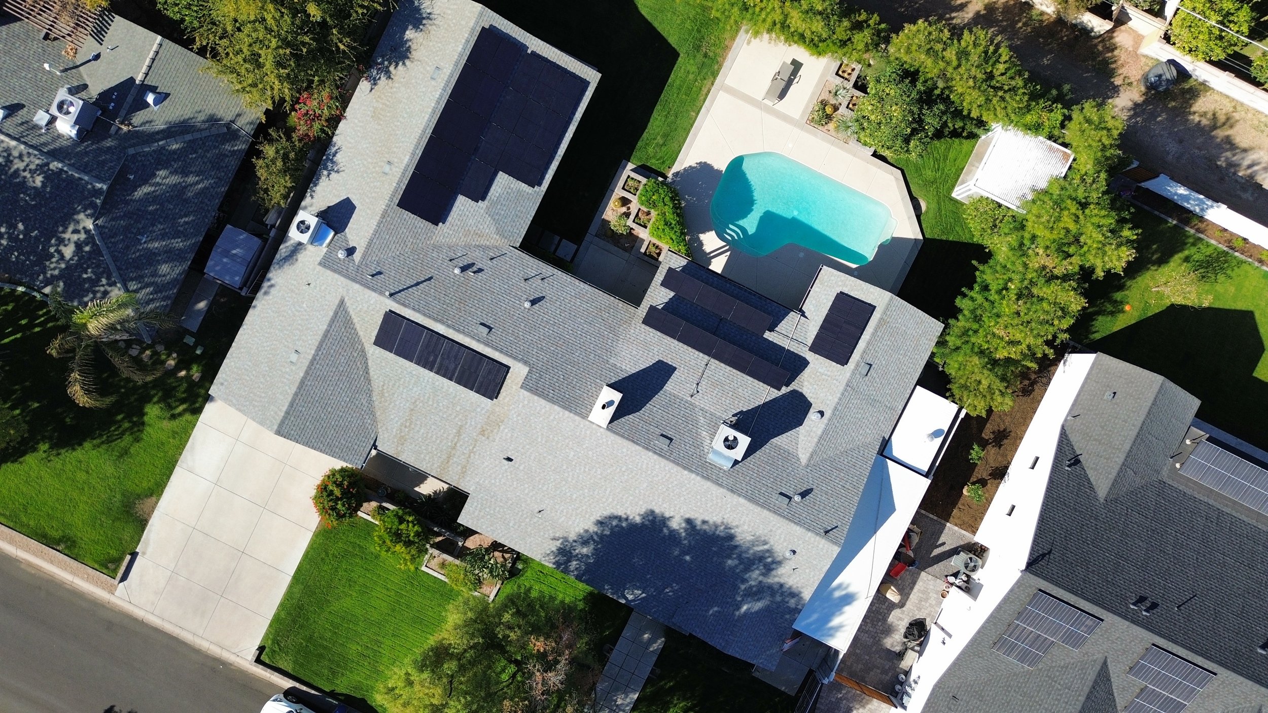 Aerial view of a residential neighborhood showing rooftops with solar panels, swimming pool, trees, lawns, and driveways.