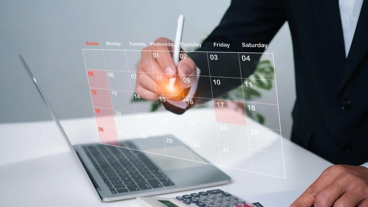 Person in business suit writing on a digital transparent calendar over a laptop and desk