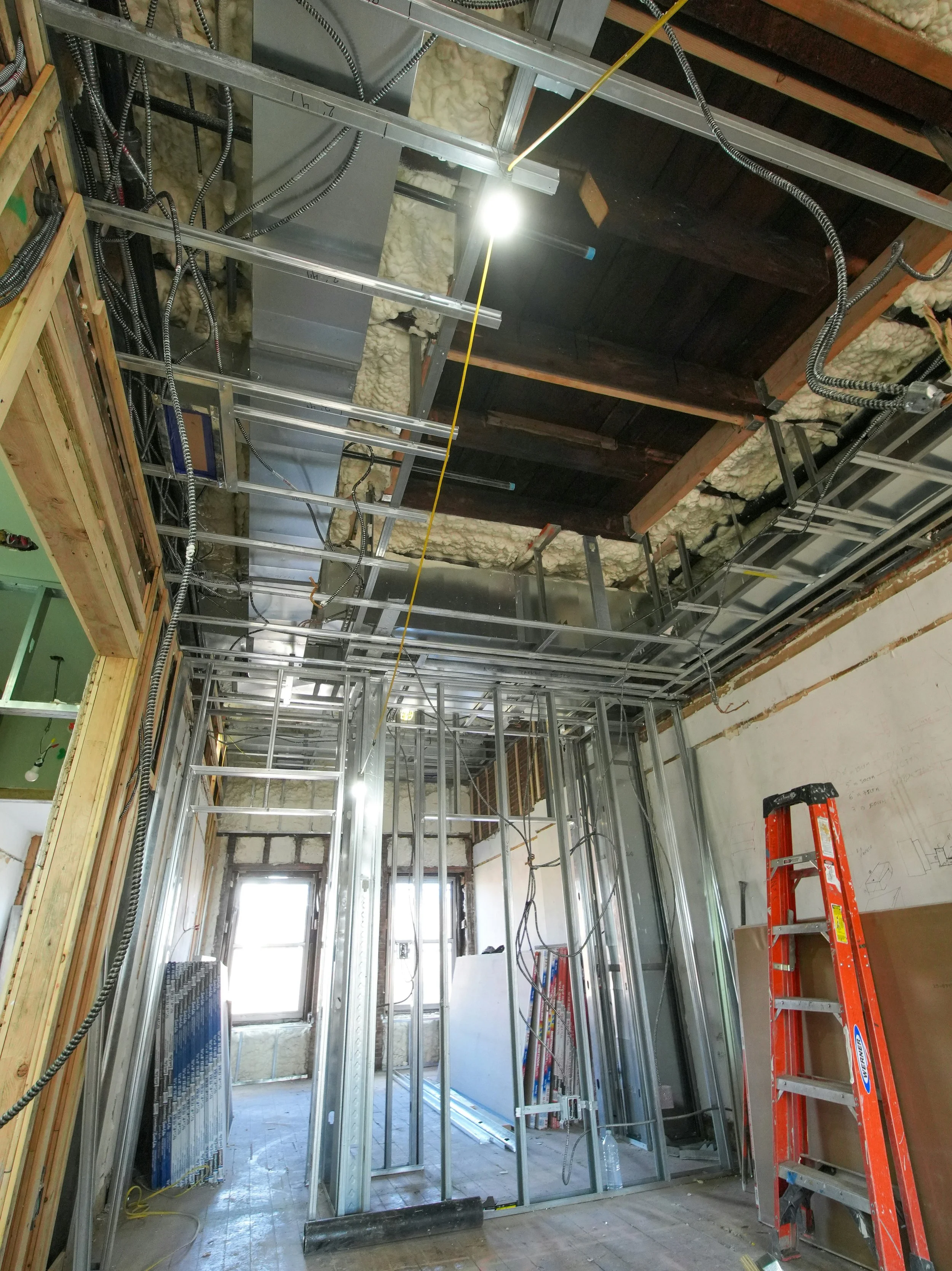 Construction site showing metal framing for interior walls, electrical wiring, insulation, and ladder, with a window at the end of the room.